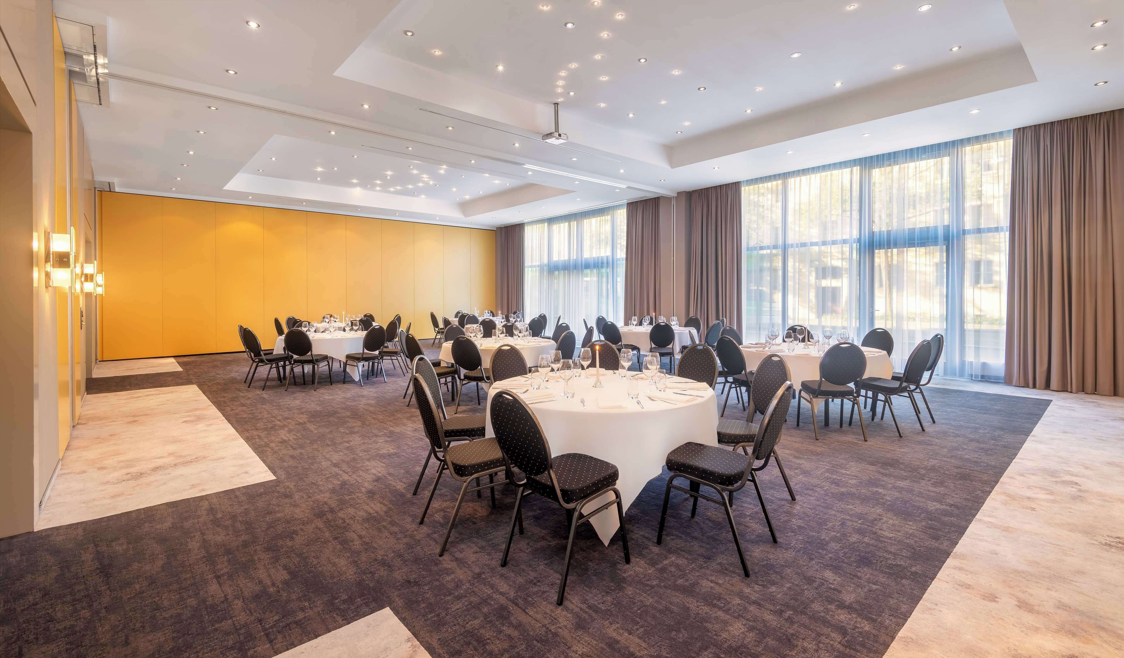 A conference room with multiple round tables, black chairs, and a large window with curtains.