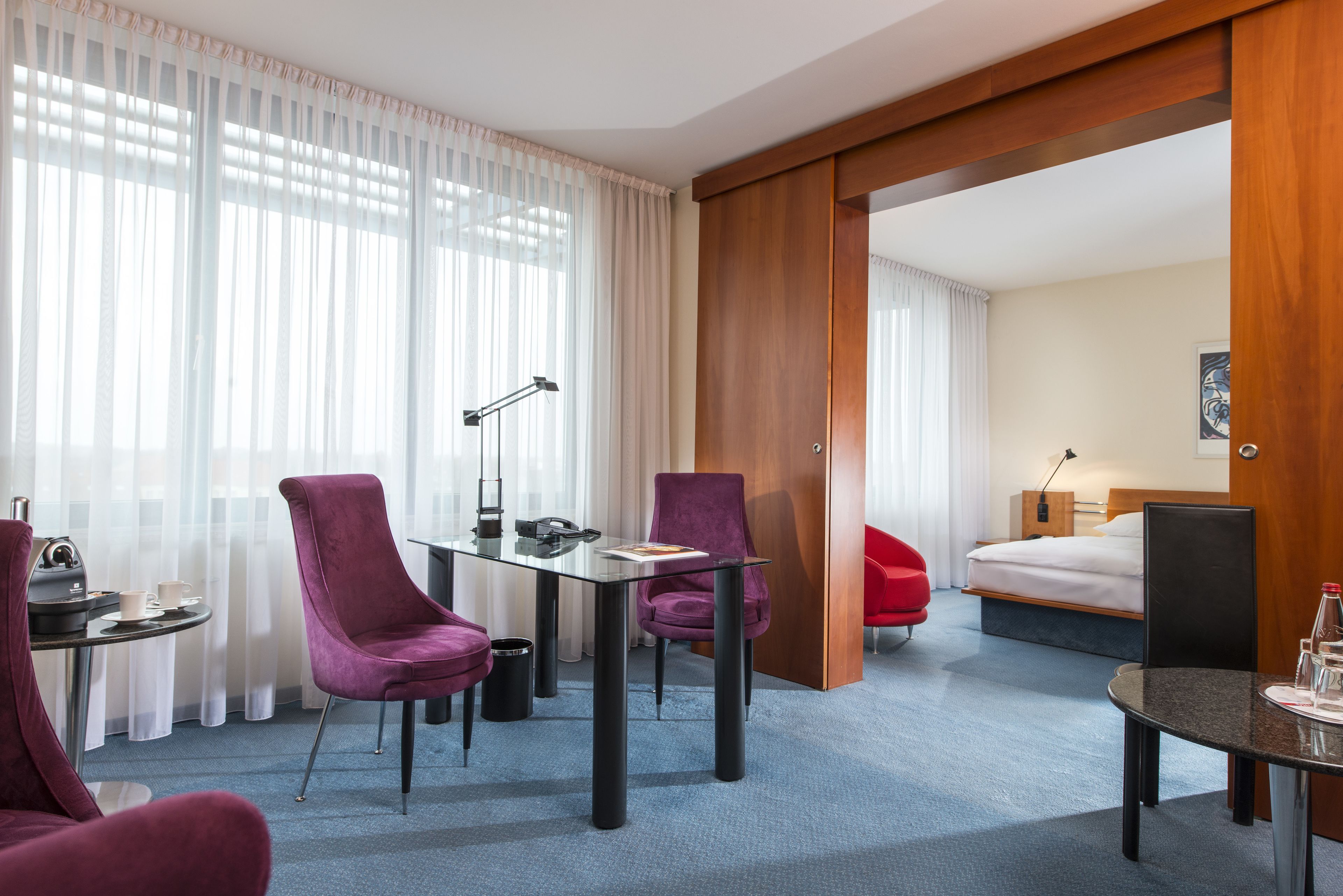 A hotel room with a bed, a desk with a lamp, two purple chairs, a small table with a cup and a phone, and a red chair.
