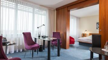 A hotel room with a bed, a desk with a lamp, two purple chairs, a small table with a cup and a phone, and a red chair.