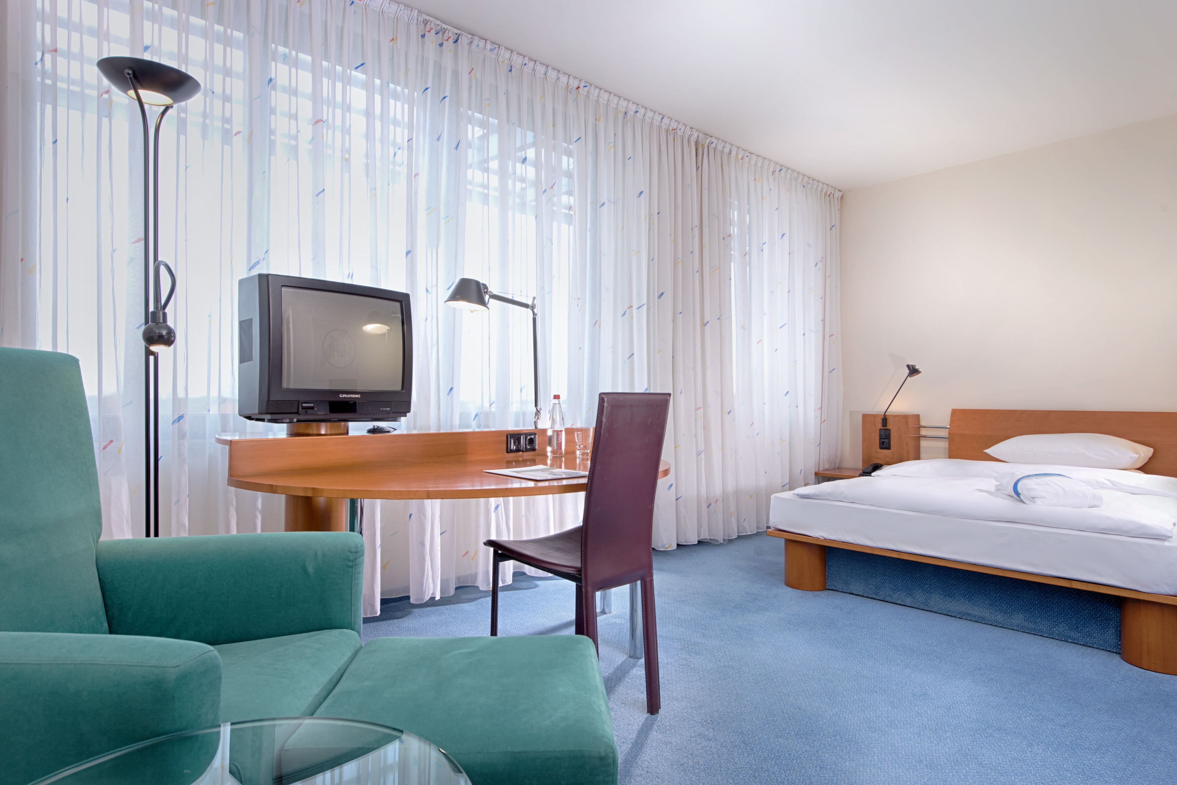 A hotel room with a bed, a desk with a television, a chair, and a lamp.