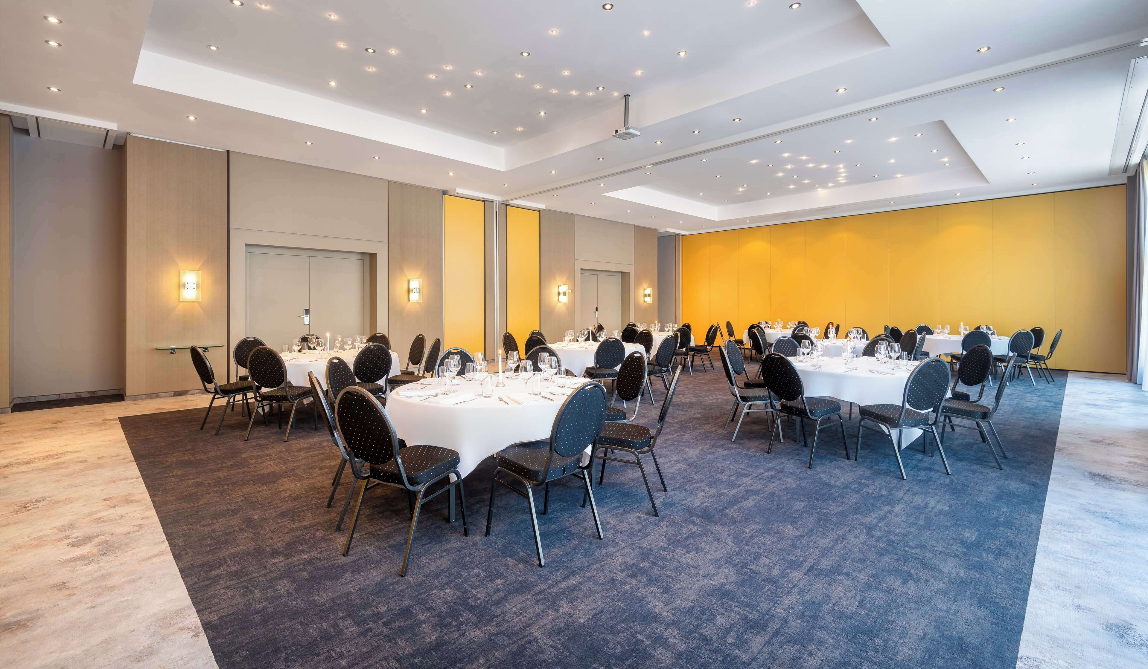 Un salón de conferencias con varias mesas redondas, sillas y piso alfombrado.