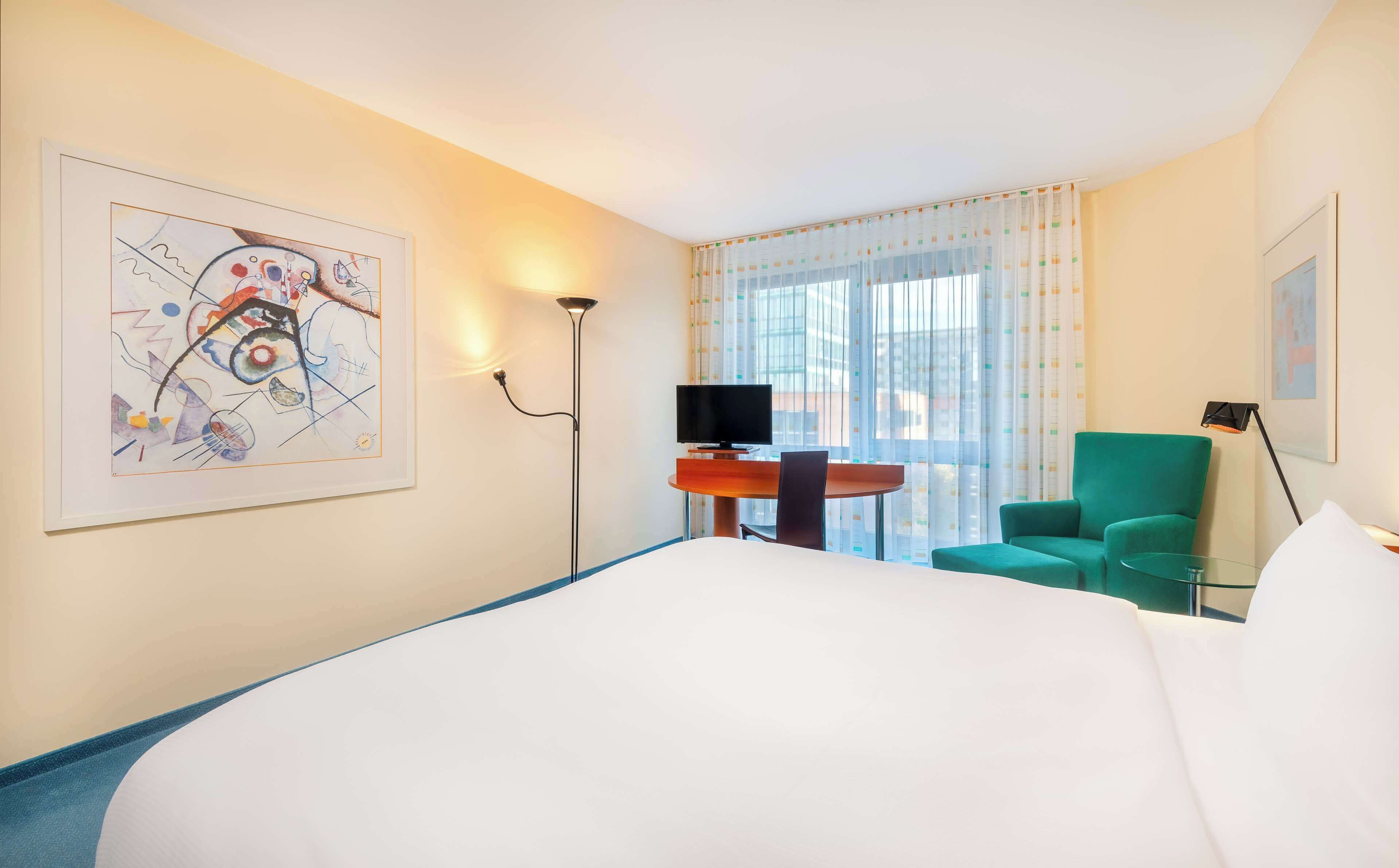 A hotel room with a large bed, a desk with a TV, a green armchair, and a painting on the wall.
