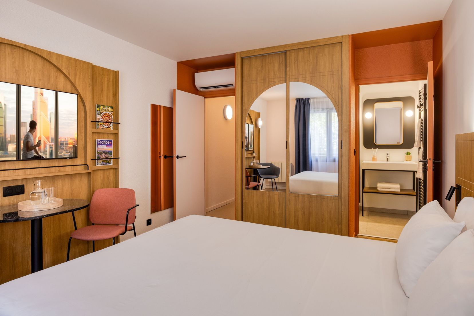Hotellihuone, jossa on sänky, työpöytä tuolilla, peili ja näkymä kaupunkiin.