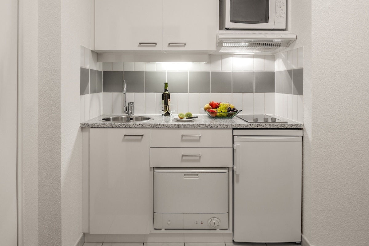 Een compacte keuken met witte kastjes, een magnetron, een spoelbak en een kleine koelkast.