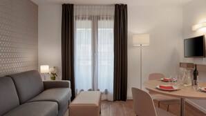 Een moderne hotelkamer met een grijze bank, een klein tafeltje met een karaf, glazen en een fles, en een aan de muur bevestigde televisie.