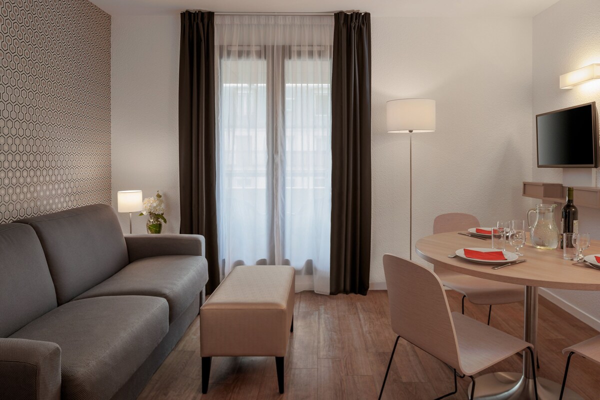 Une chambre d’hôtel moderne avec un canapé gris, une petite table sur laquelle se trouvent une carafe, des verres et une bouteille, et un téléviseur fixé au mur.