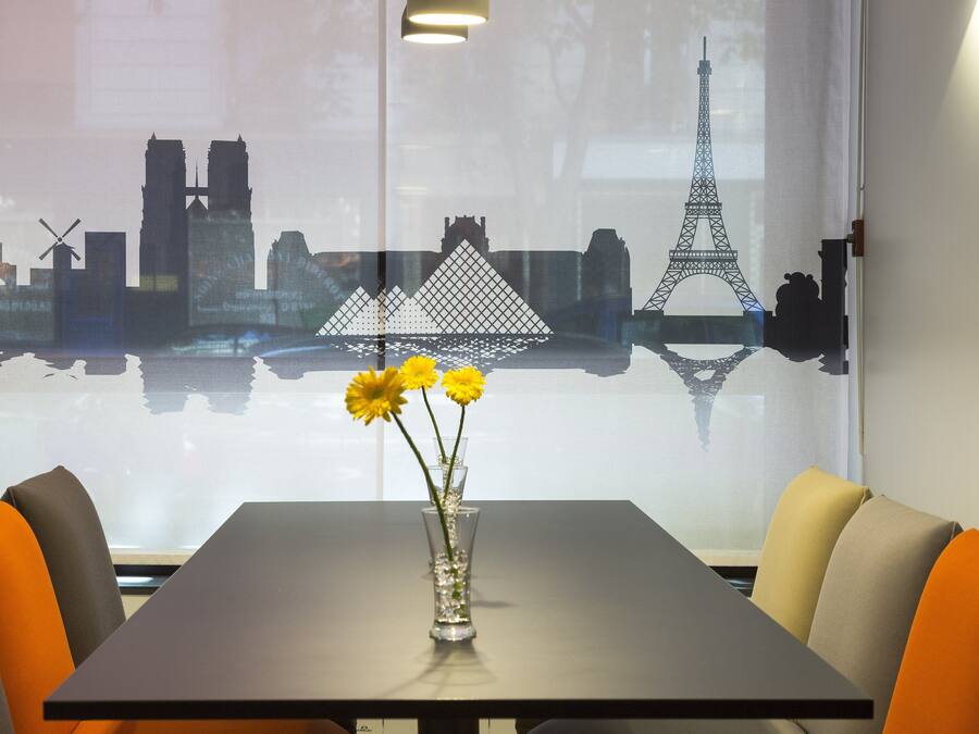 Una mesa con un jarrón de flores amarillas, vista panorámica de la ciudad y asientos naranja y grises.