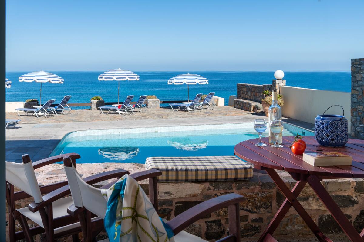 Een zwembadgedeelte met ligbedden en parasols, een tafel met een fles, glazen en een boek, en uitzicht op de oceaan.