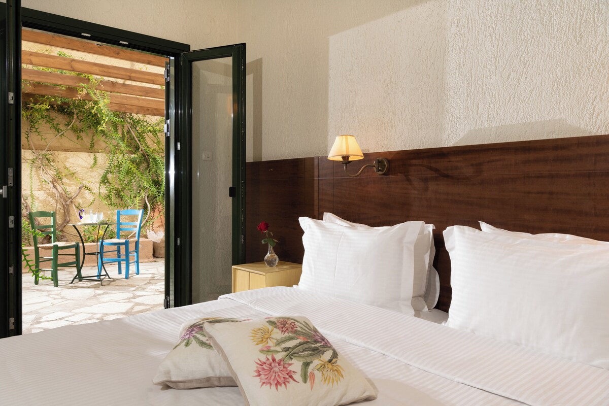 Une chambre d’hôtel avec un lit, une table de chevet, une lampe et une vue sur une terrasse extérieure avec des chaises et une table.