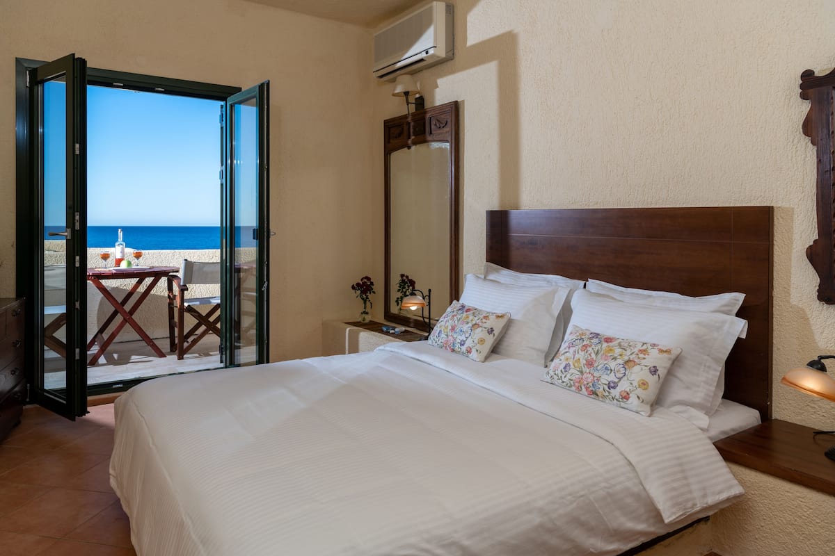 Een hotelkamer met een groot bed, een nachtkastje, een spiegel en uitzicht op het strand door een open deur.