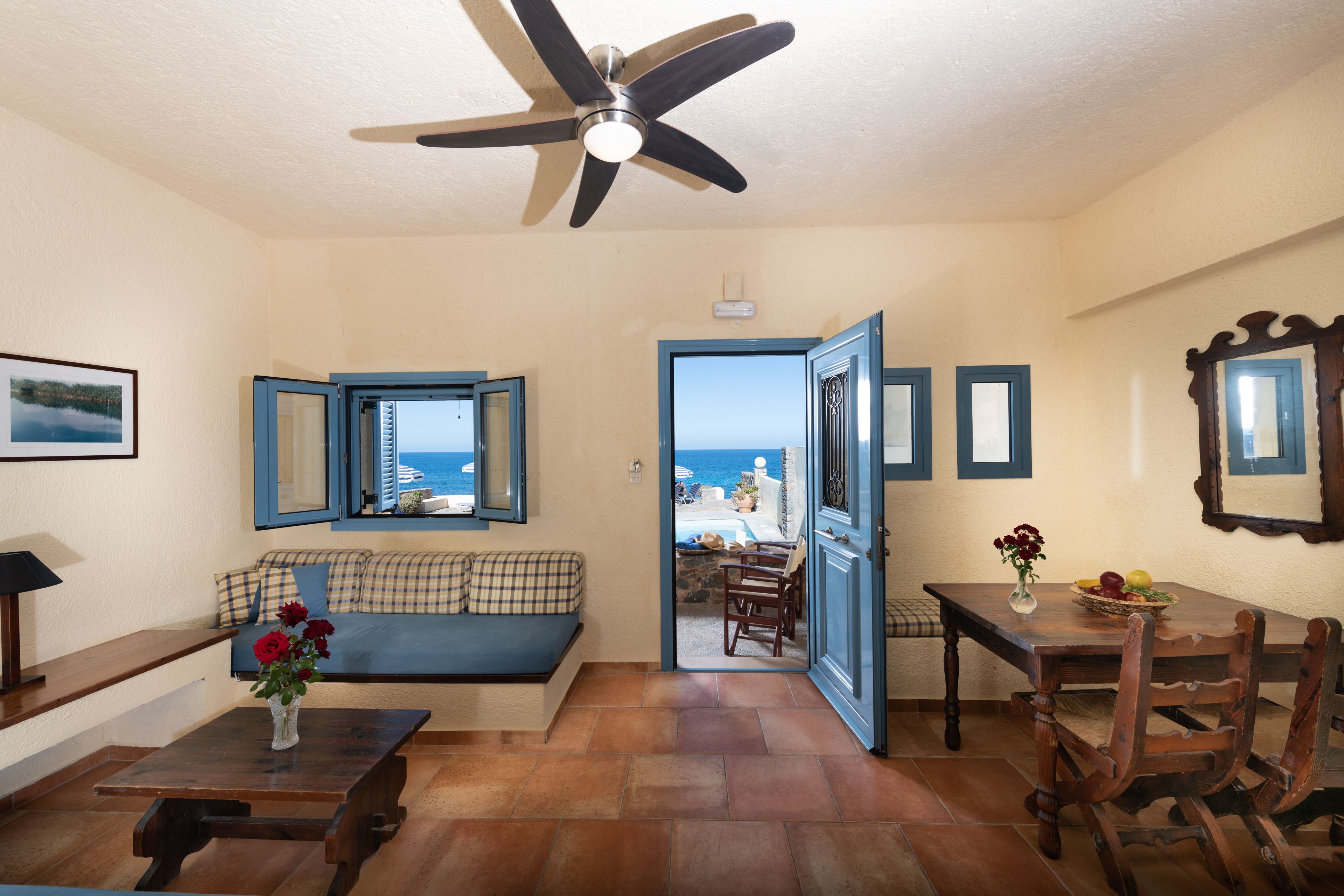 A room with a ceiling fan, a sofa, a dining table with chairs, a vase with flowers, and a view of the sea through a window.