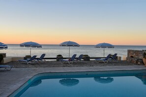 Un espace aménagé au bord de la piscine, avec des chaises longues et des parasols, un mur en pierre et une vue sur la mer au coucher du soleil.