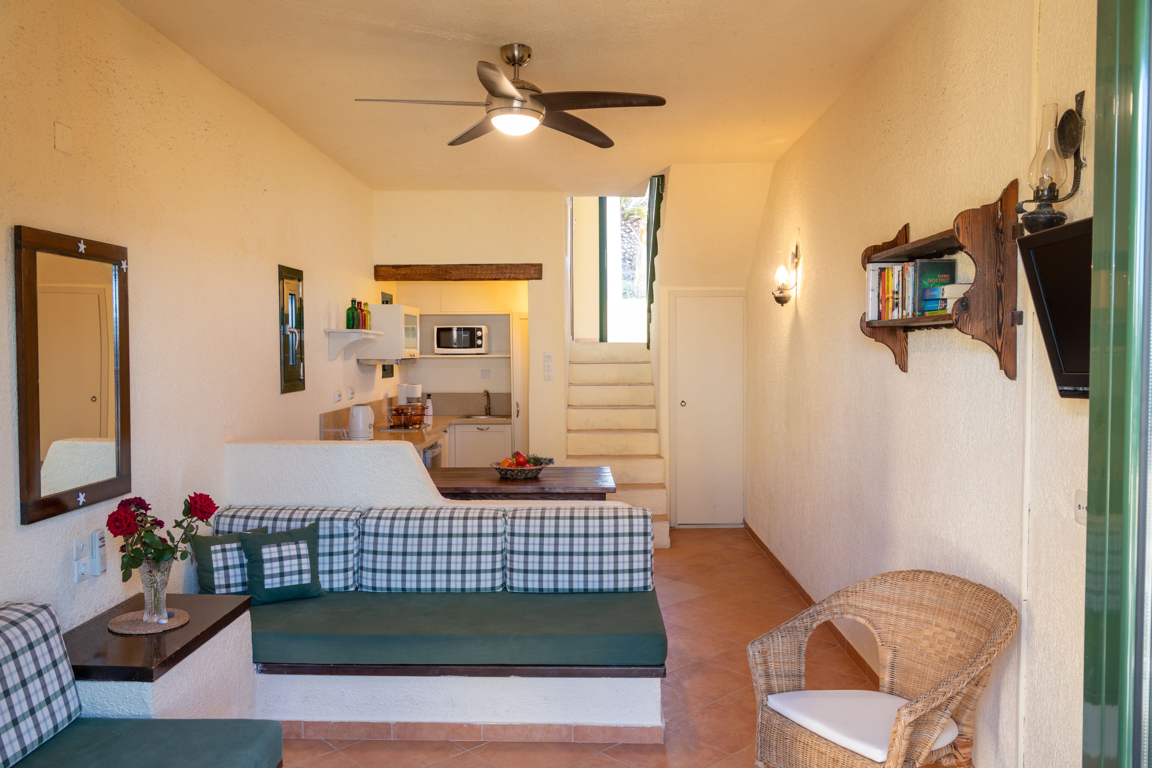 A compact living space with a kitchenette, a ceiling fan, a sofa, a wicker chair, and a bookshelf.