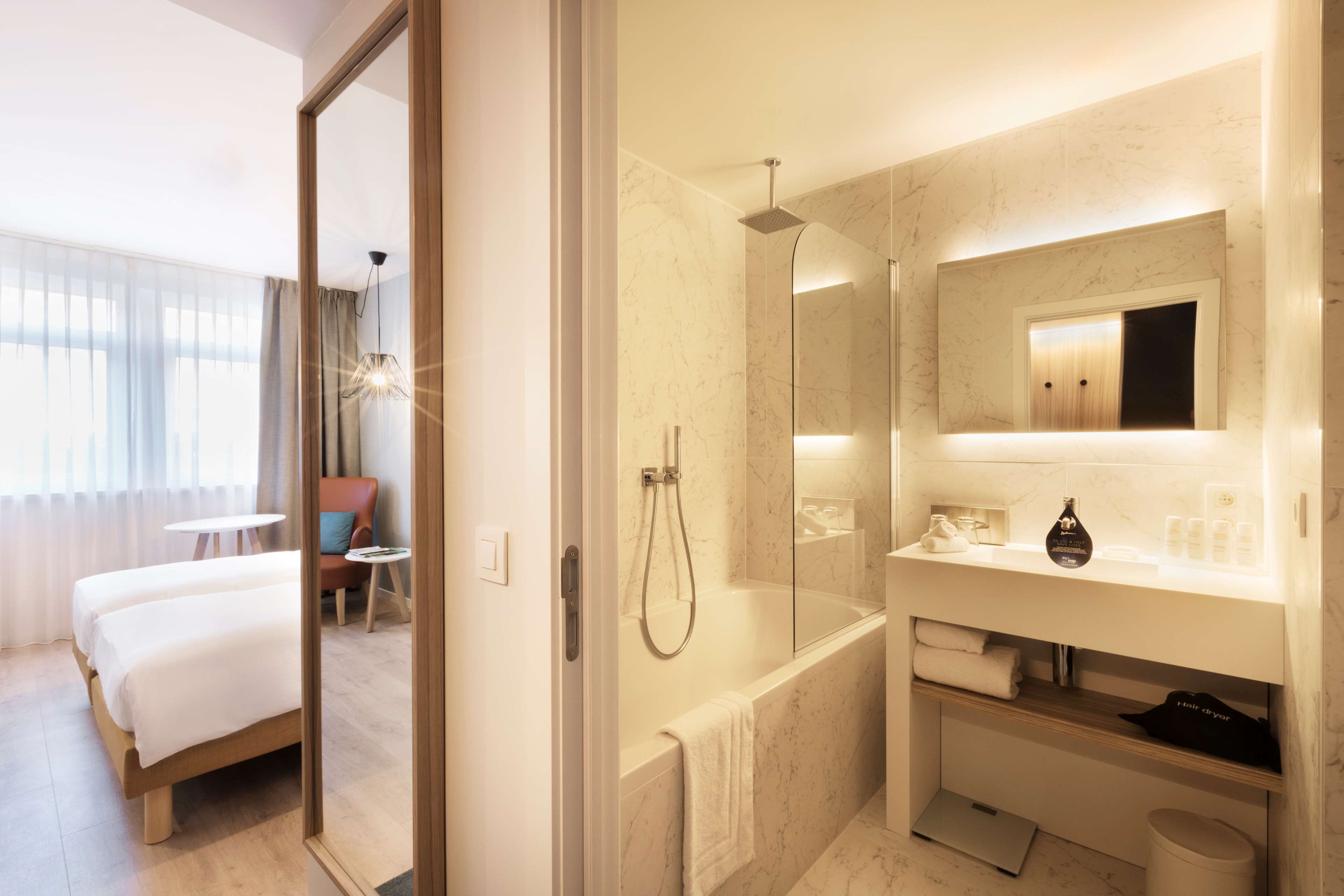 A modern hotel room with a large bed, a separate bathroom with a glass shower enclosure, and a small seating area with a chair and table.