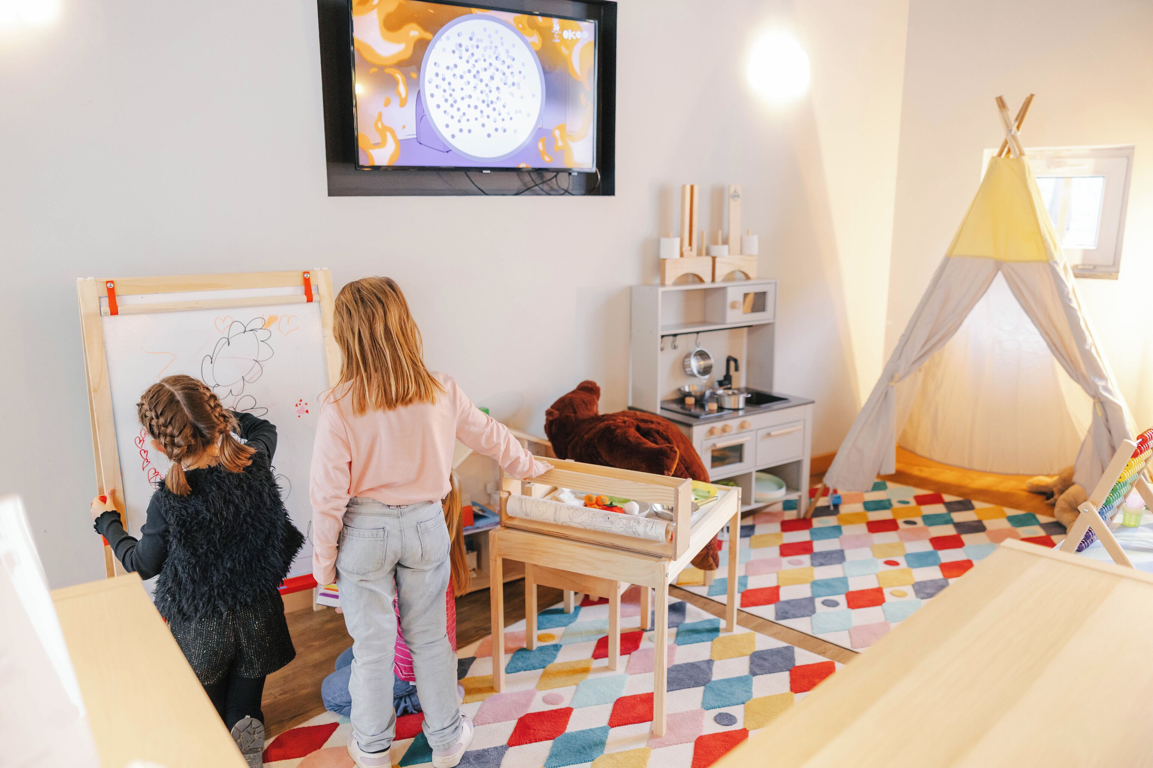 A child drawing on a whiteboard, a child playing with a toy, and a child sitting on a chair in a room with a colorful floor, a television, and a tent-like structure.