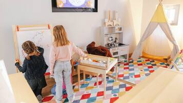 A child drawing on a whiteboard, a child playing with a toy, and a child sitting on a chair in a room with a colorful floor, a television, and a tent-like structure.