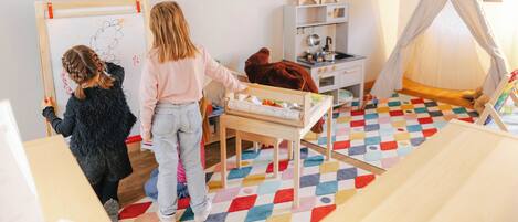 Uma criança desenhando em um quadro branco, uma criança brincando com um brinquedo e uma criança sentada em uma cadeira em um quarto com piso colorido, uma televisão e uma estrutura parecida com uma tenda.