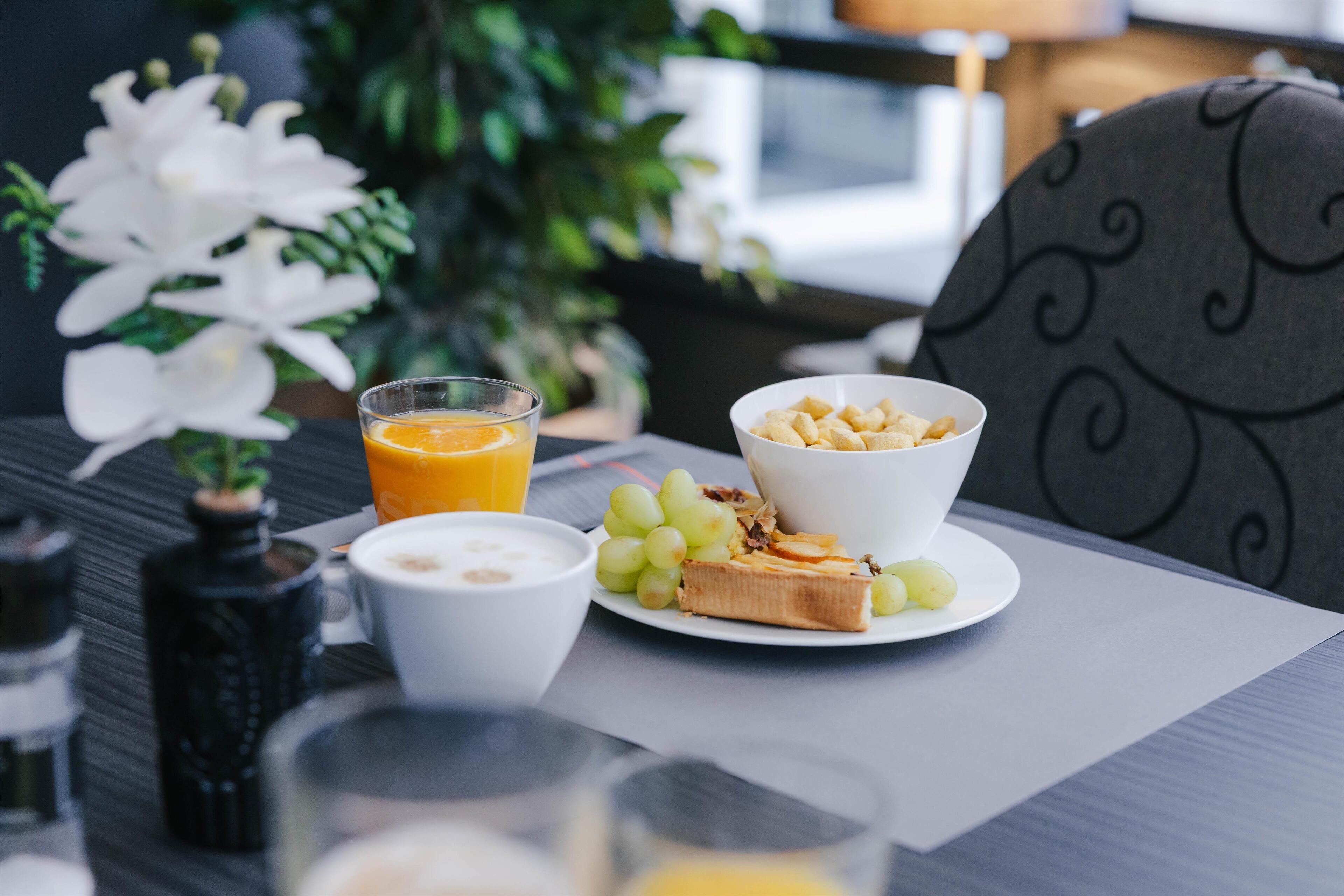 A breakfast setting with a plate of toast, grapes, and a bowl of cereal, a glass of orange juice, and a cup of coffee on a table.
