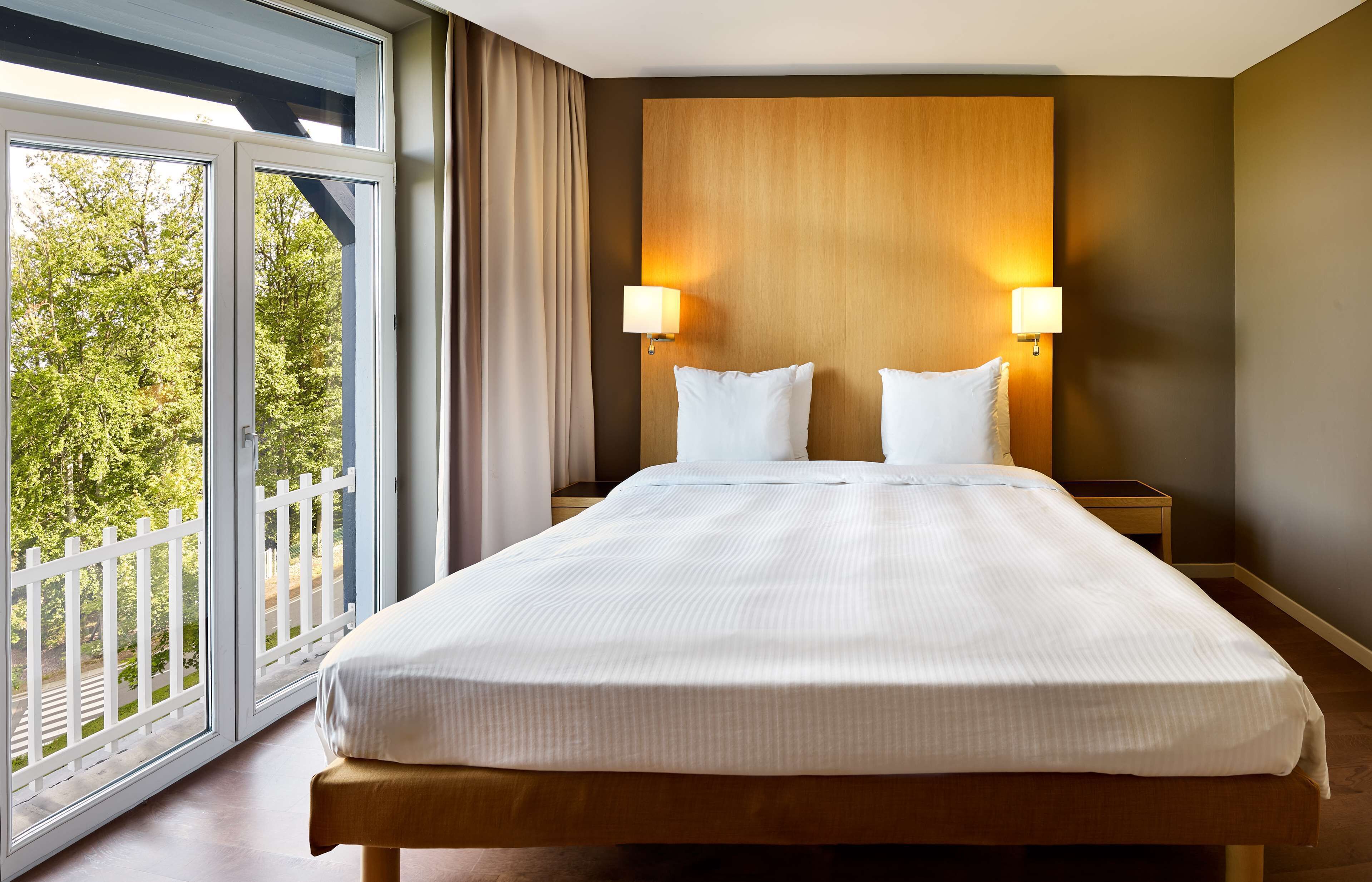 A hotel room with a large bed, two bedside lamps, a wooden headboard, and a view of trees through large windows.