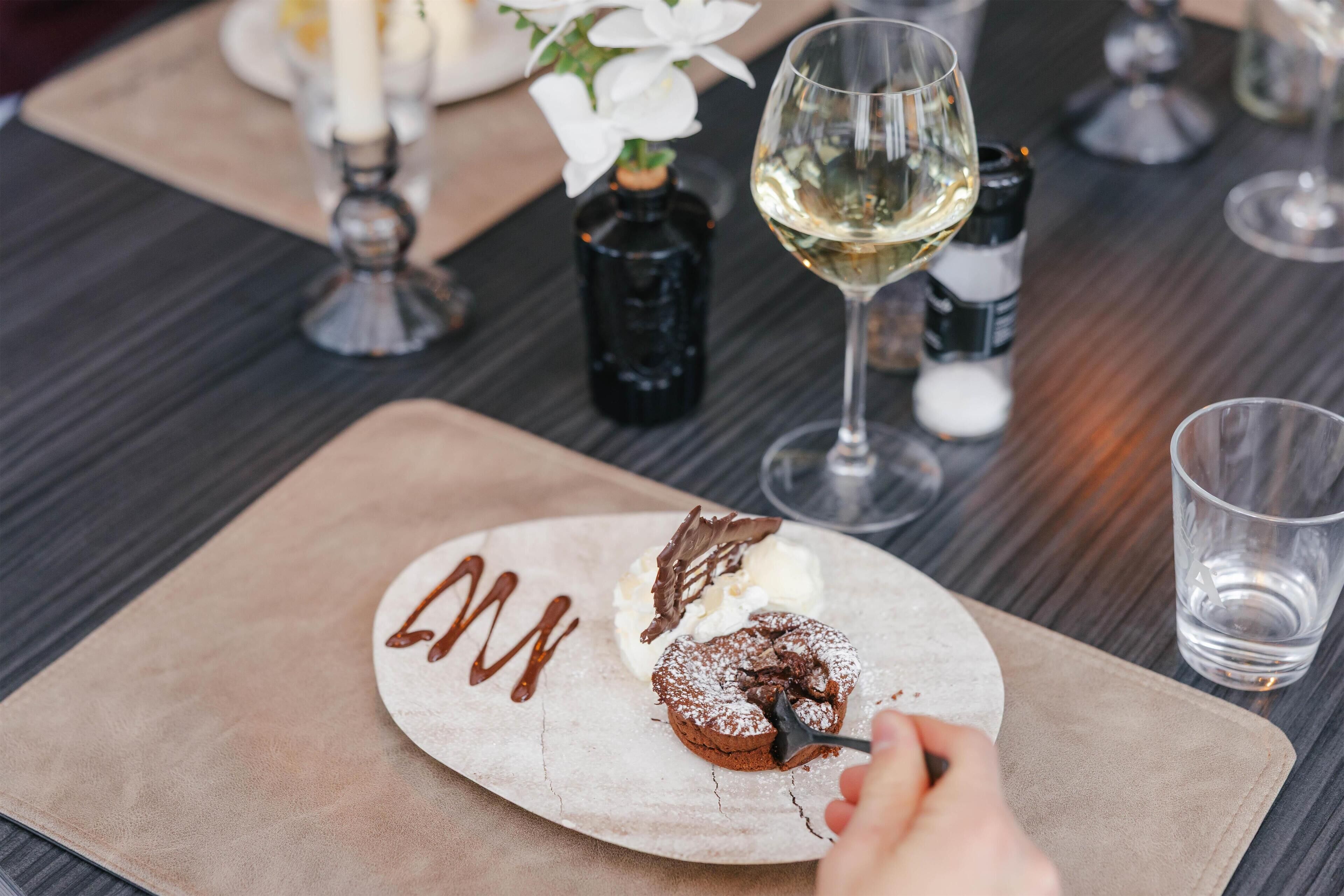 A dessert plate with a chocolate cake and ice cream, a glass of white wine, and a vase with white flowers on a table.