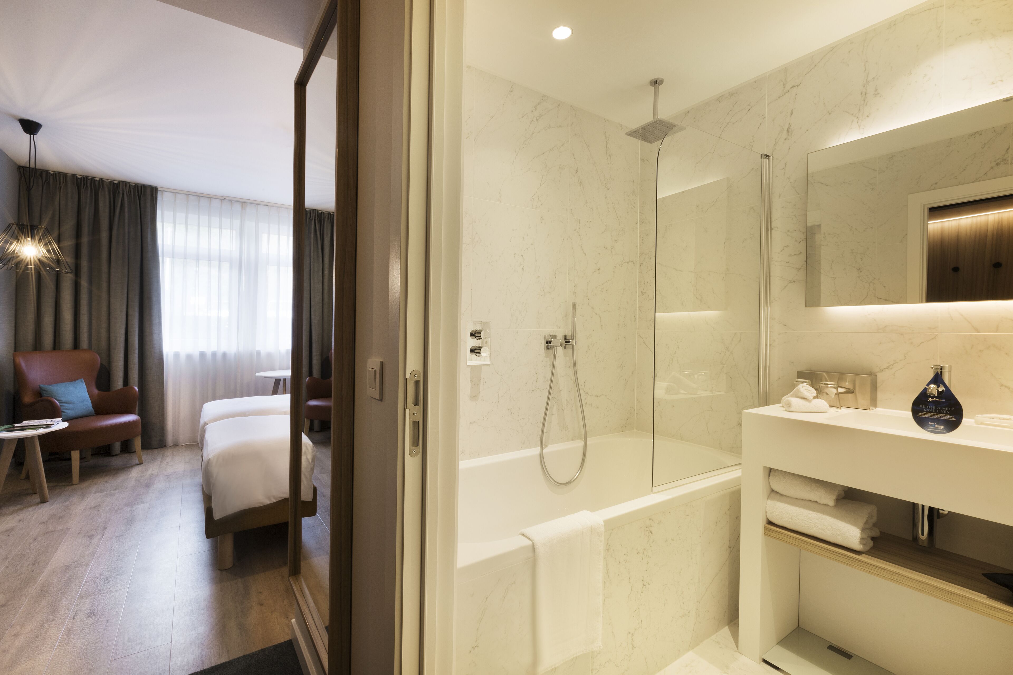 A modern hotel room with a large bed, a sitting area with a chair and table, a bathroom with a bathtub and shower, and a vanity with a mirror and towel rack.