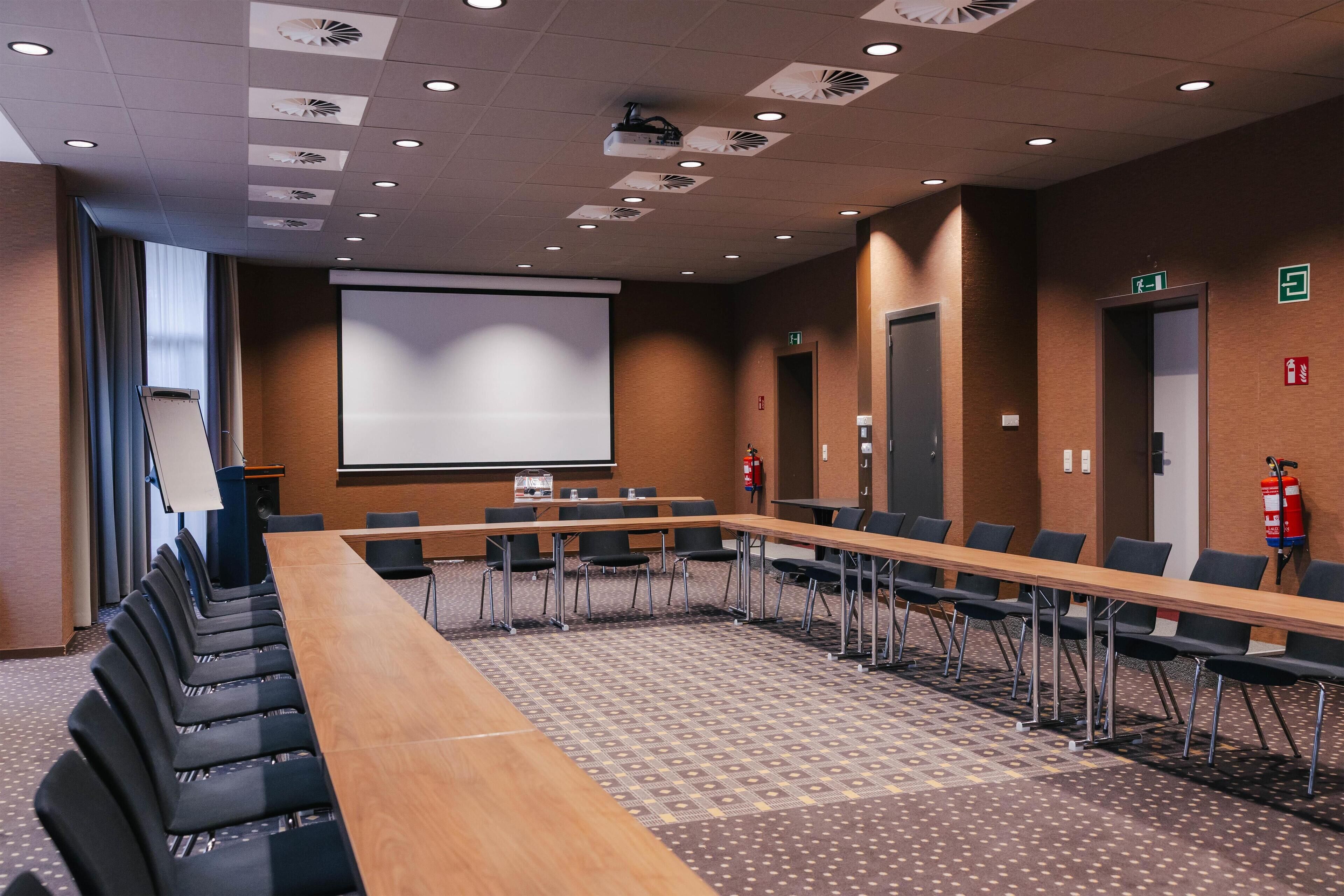 Un salón de conferencias con una larga mesa, sillas, una pantalla de proyector y un extintor.