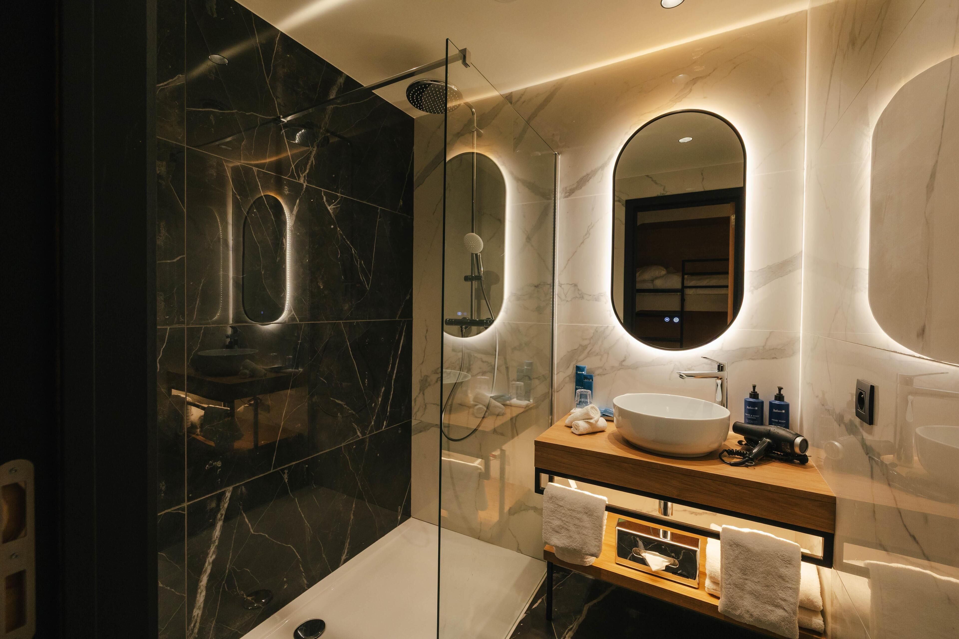 A modern bathroom with a glass-enclosed shower, a marble-tiled wall, a wooden vanity with a large sink, and a mirror above the vanity.