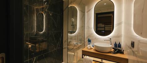 Un baño moderno con cabina de ducha acristalada, pared con azulejos de mármol, tocador de madera con un lavamanos grande y un espejo sobre el tocador.