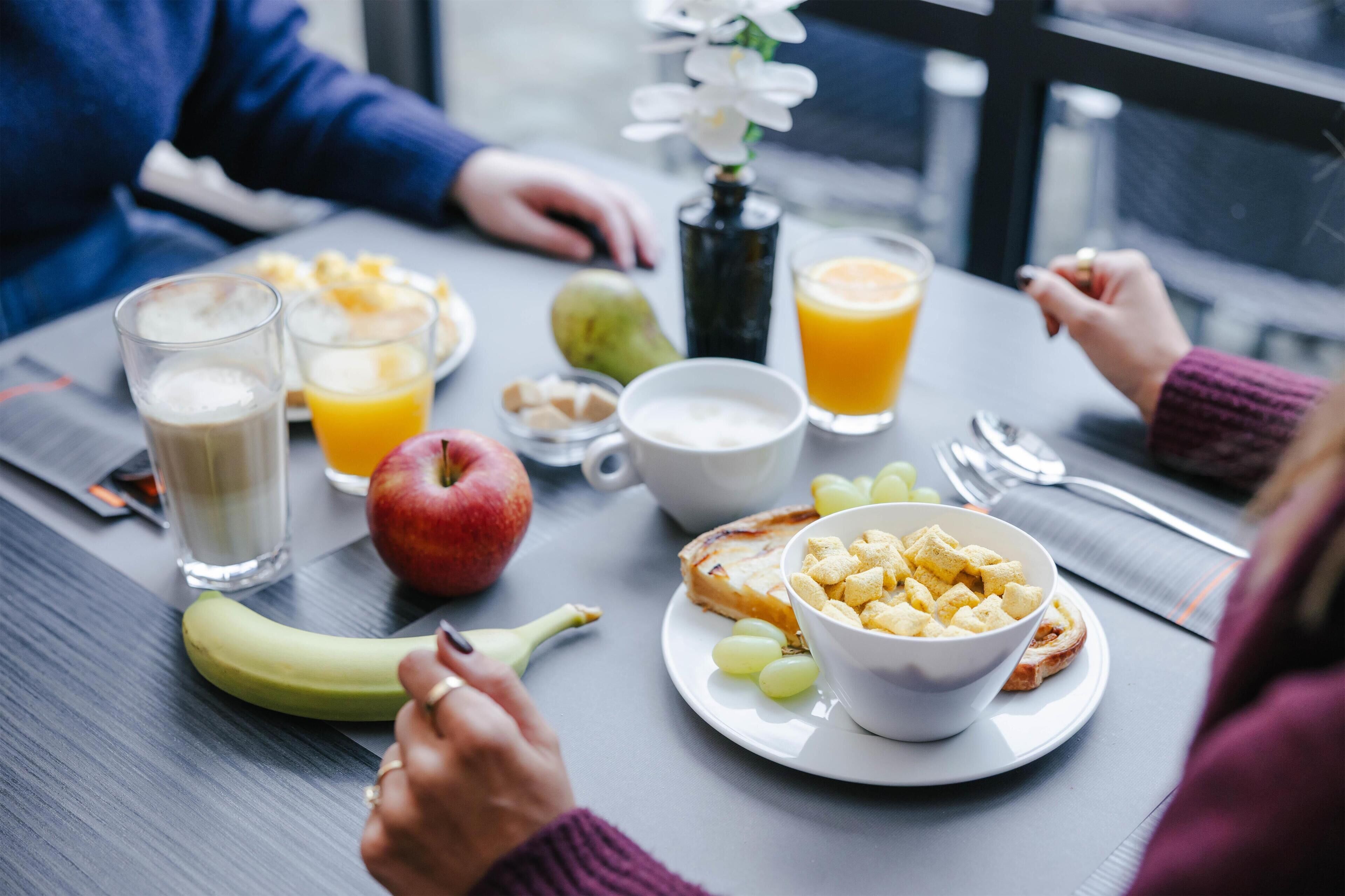 A breakfast table with various food items and drinks, including a banana, apple, toast, cereal, and orange juice.