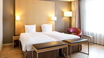 A hotel room with two beds, a wooden headboard, bedside lamps, a red chair, a small table, and a large window with curtains.