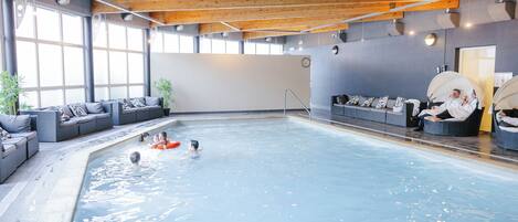 Una piscina interior con techo de madera, ventanas amplias y zonas de estar con sombrillas.