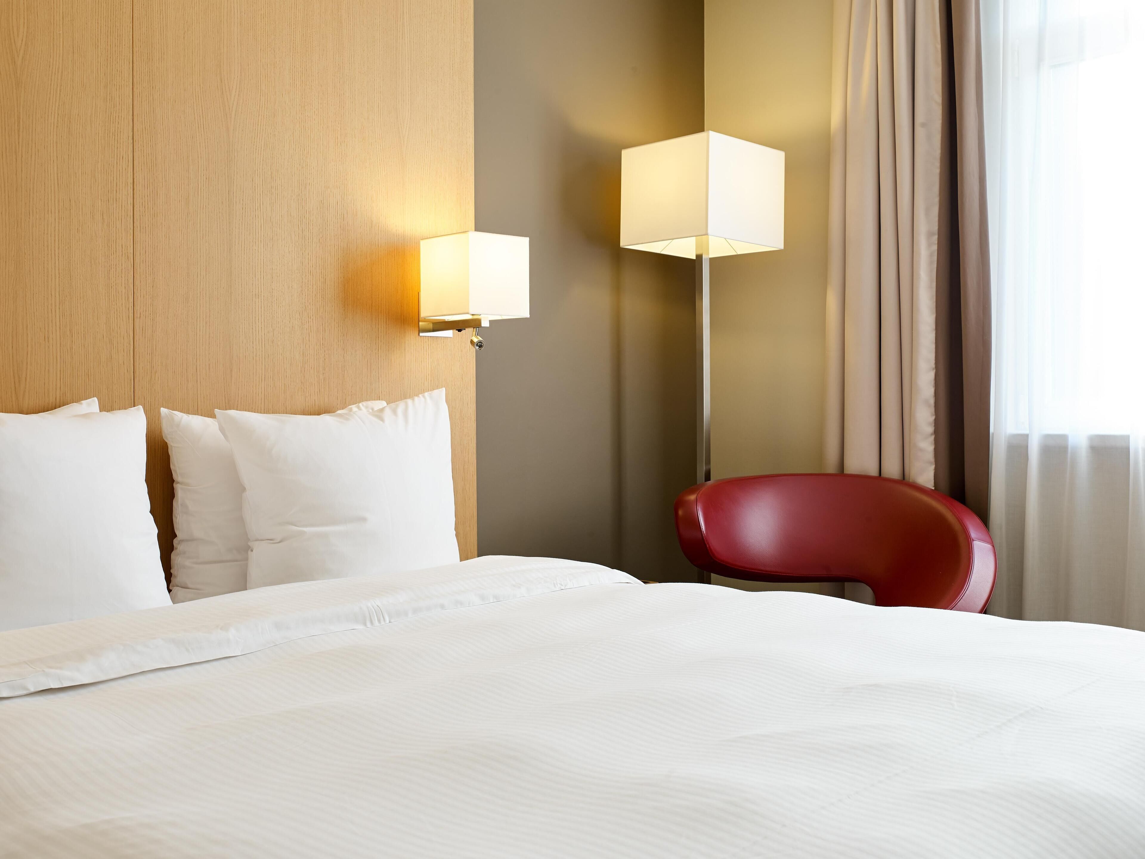 Habitación de hotel con cama, una silla roja, una lámpara y vistas al exterior.