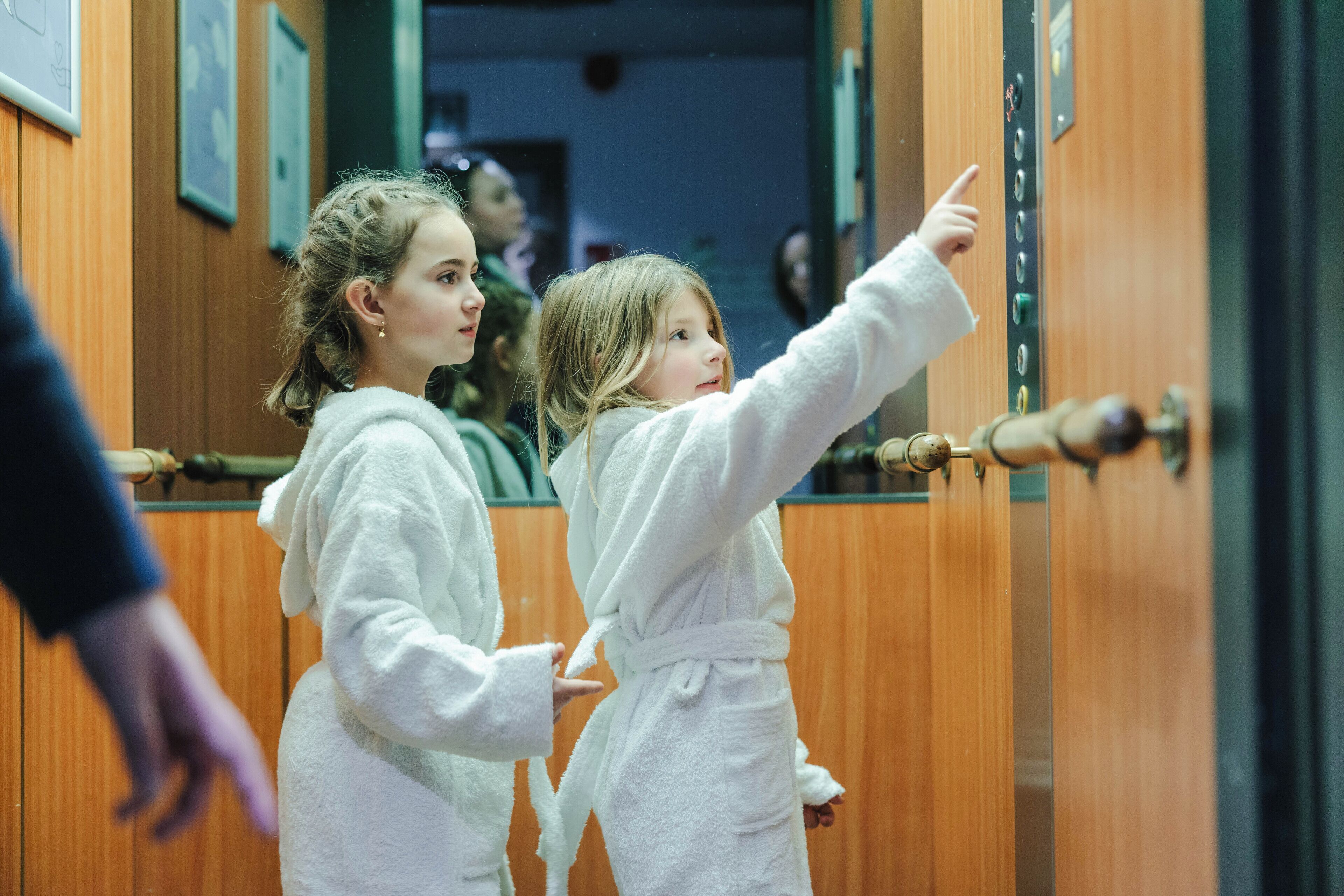 Two individuals in white robes standing in a hallway with wooden paneling and a mirror on the wall.