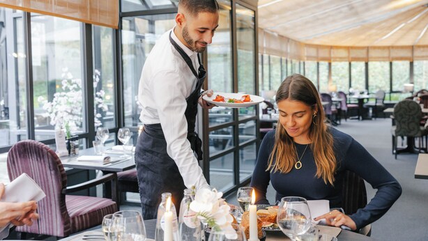 Un restaurante con un interior moderno, ventanas amplias y un camarero sirviendo comida a un cliente.
