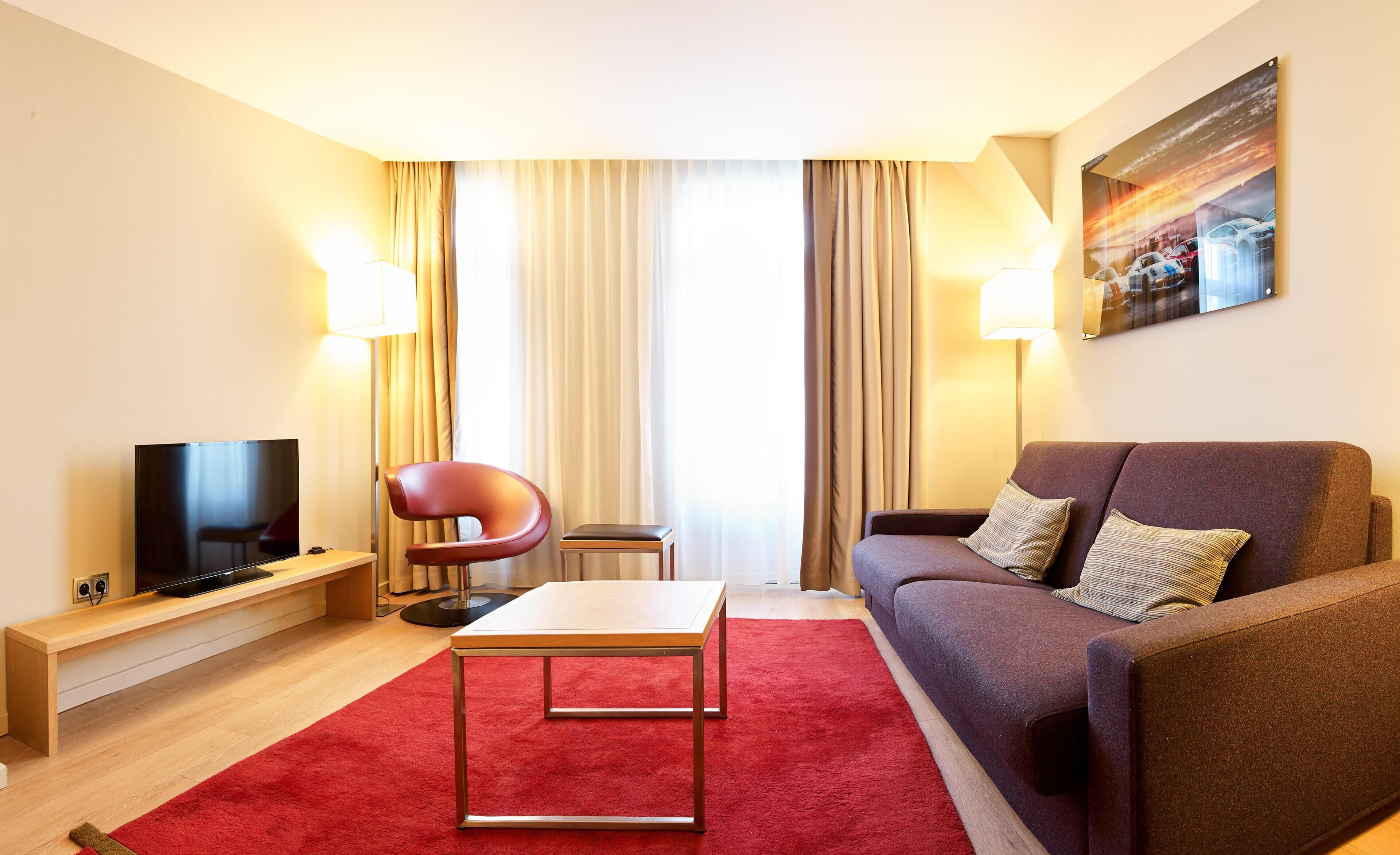 A modern hotel room with a flat-screen TV, a red armchair, a brown sofa, a small table, and a red carpet.