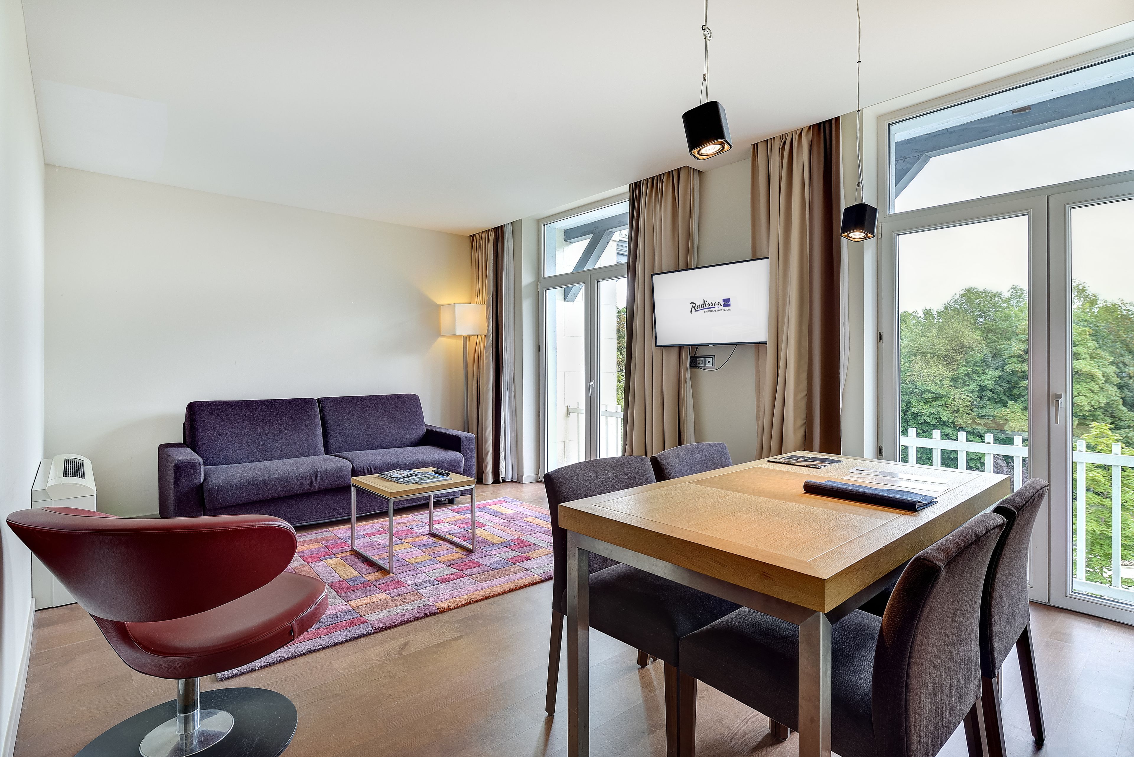 A modern hotel room with a wooden dining table, a red chair, a sofa, and a TV mounted on the wall.