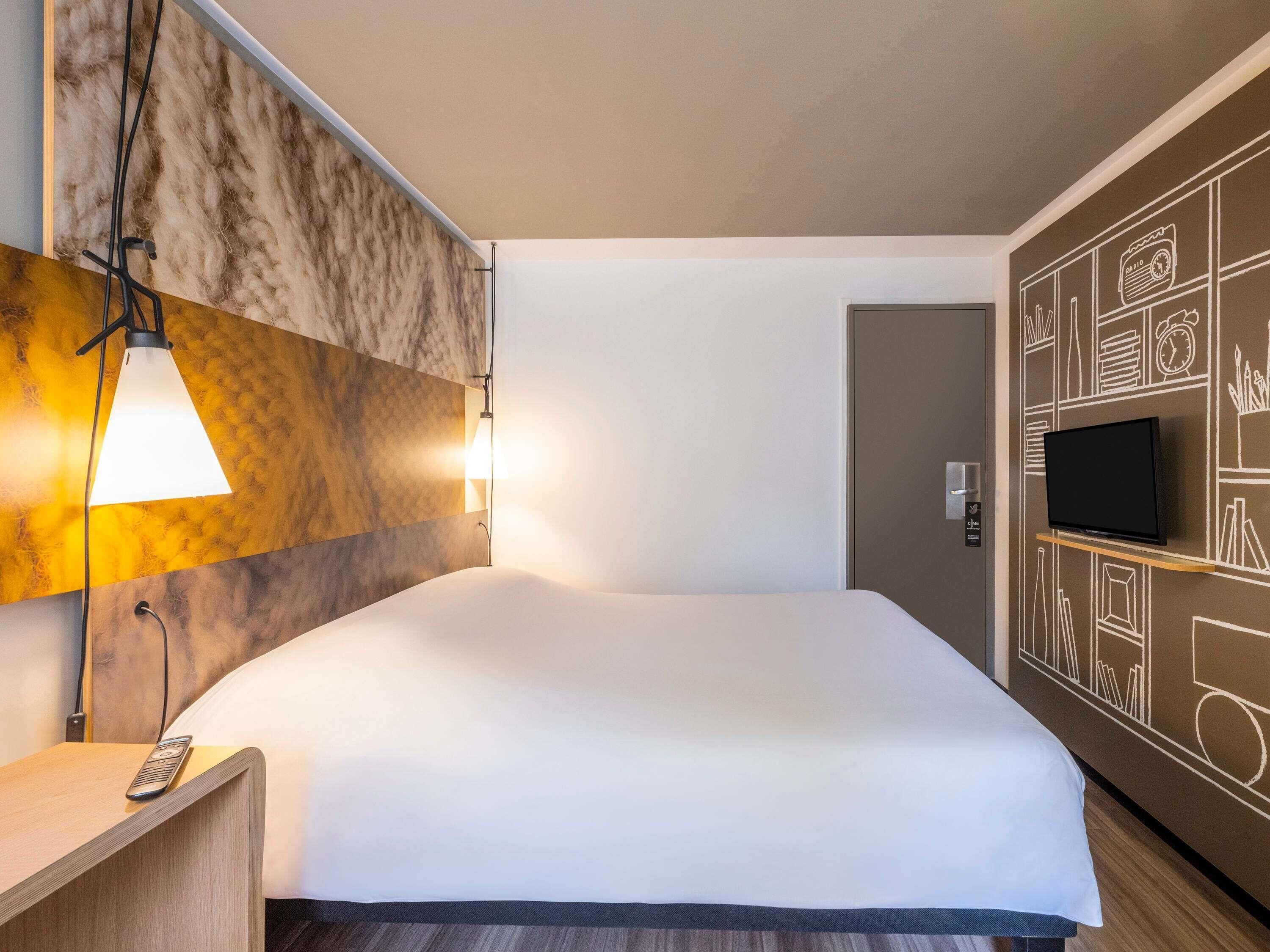 Ein Hotelzimmer mit einem großen Bett, einem Nachttisch mit Fernbedienung, einem an der Wand montierten Fernseher und einem einzigartigen Wanddesign mit geometrischen Mustern.