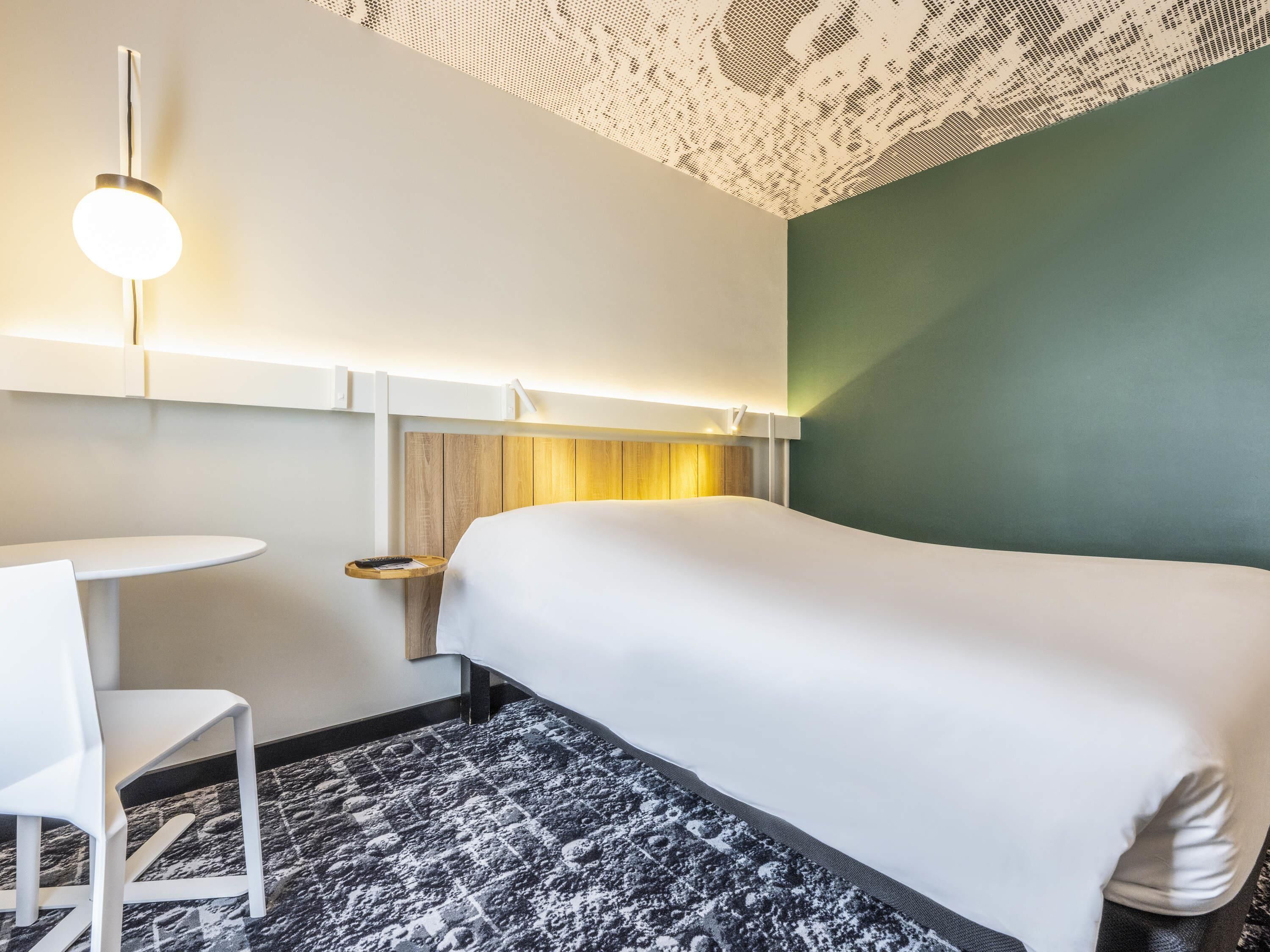 A hotel room with a bed, a small table, a chair, and a wall-mounted light fixture.
