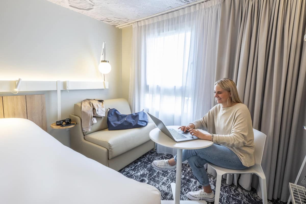 Ein Hotelzimmer mit Bett, einer Couch, einem kleinen runden Tisch mit einem Laptop, einer Lampe und einem Fenster mit Vorhängen.