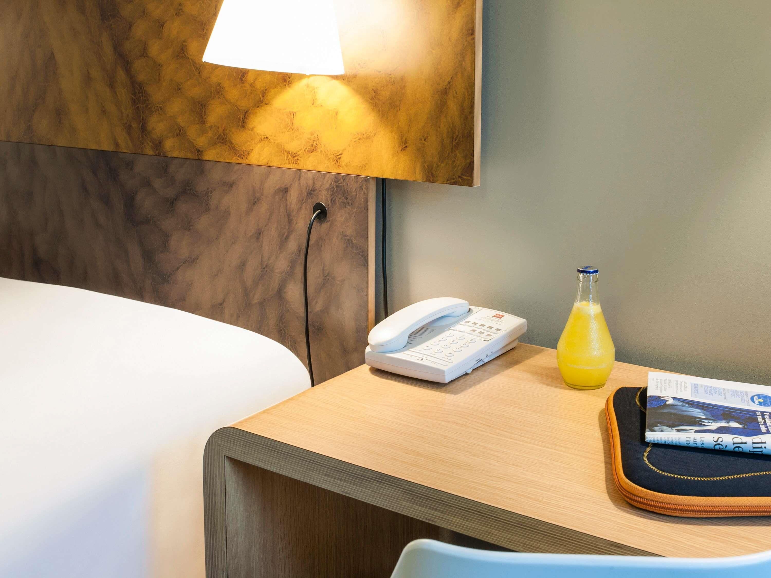 A hotel room with a bed, a bedside table with a lamp, a phone, a glass of orange juice, and a magazine.