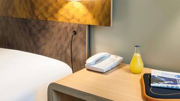 A hotel room with a bed, a bedside table with a lamp, a phone, a glass of orange juice, and a magazine.