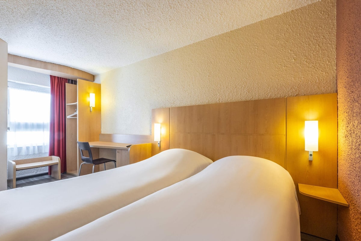 Ein Hotelzimmer mit zwei Betten, einem Schreibtisch, einem Stuhl und einem Fenster mit Vorhängen.