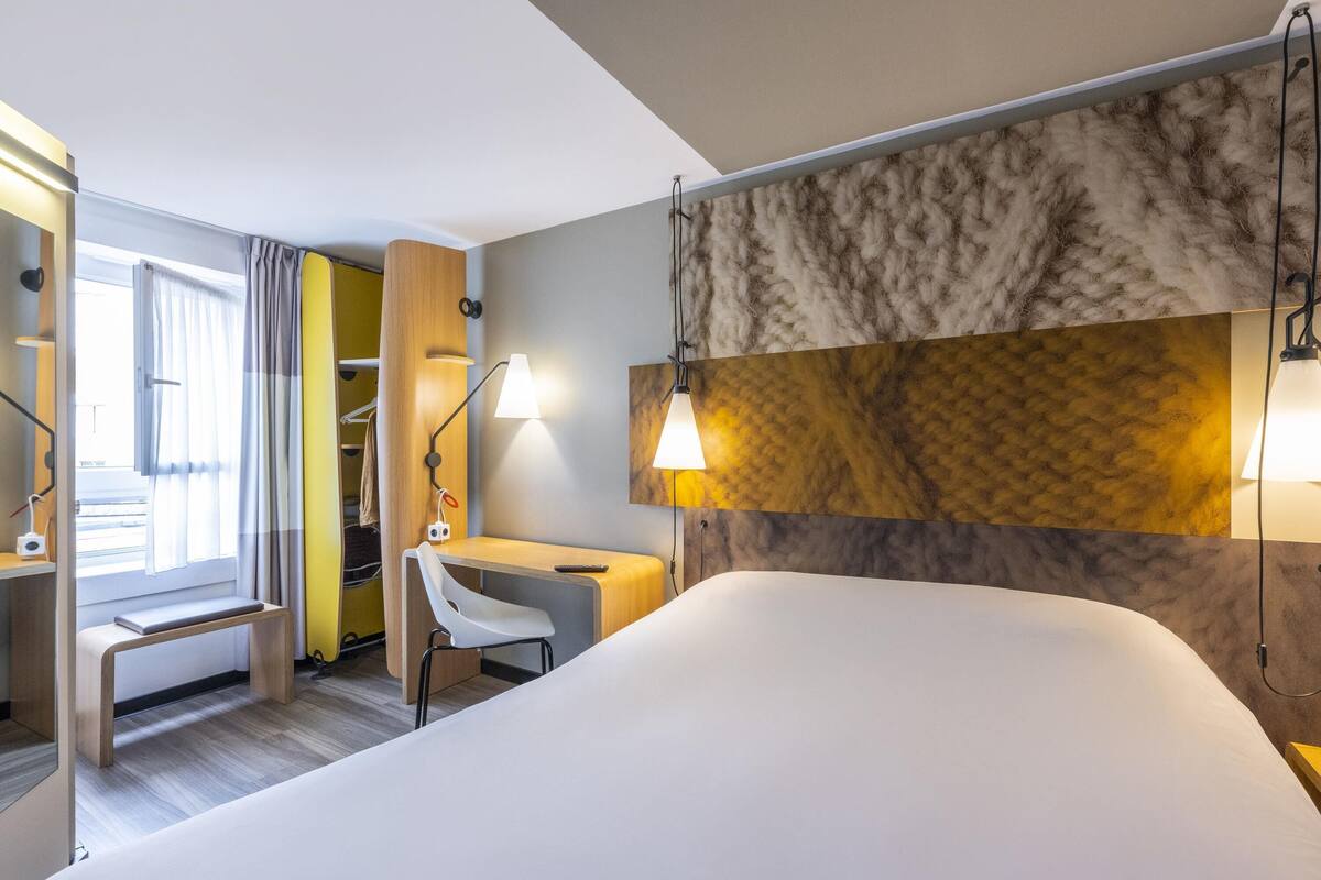 Ein Hotelzimmer mit einem Bett, einem Schreibtisch, einem Stuhl, einem Kleiderschrank und einem Fenster mit Vorhängen.