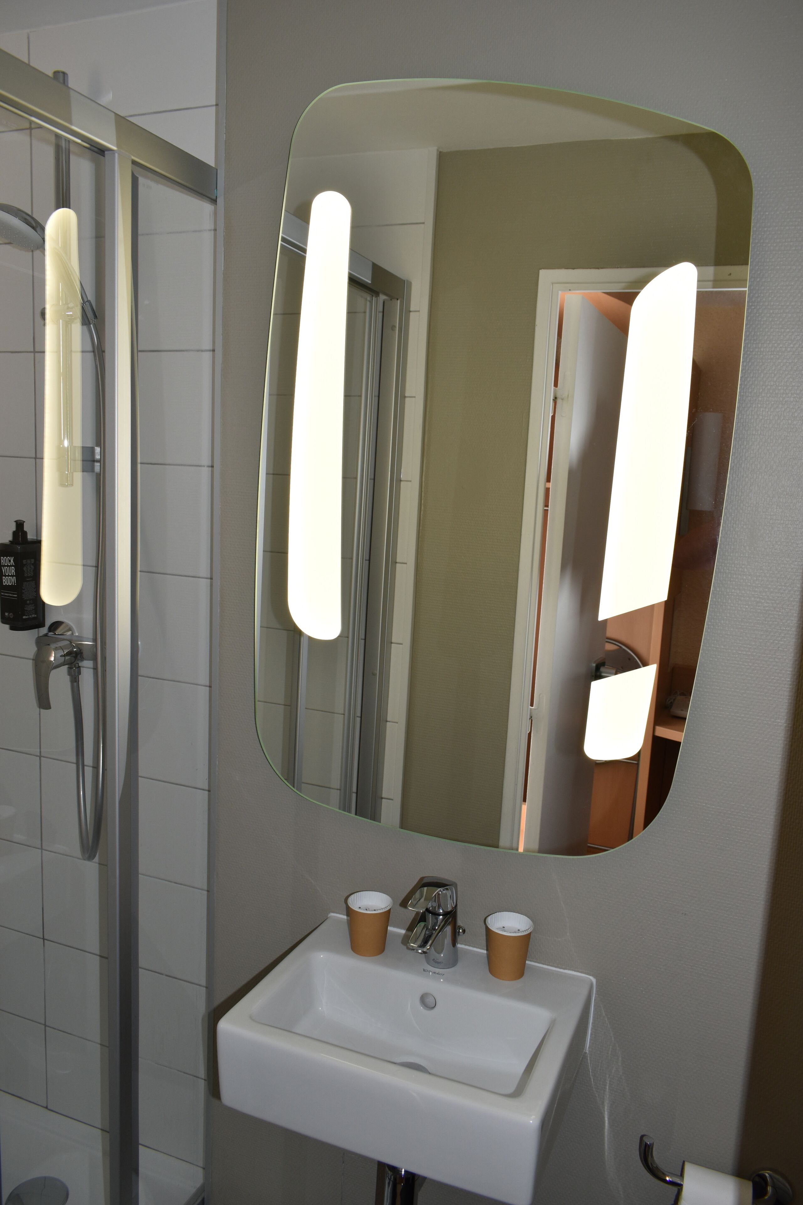 Un baño con lavamanos blanco, dos tazas, un espejo y zona de ducha.