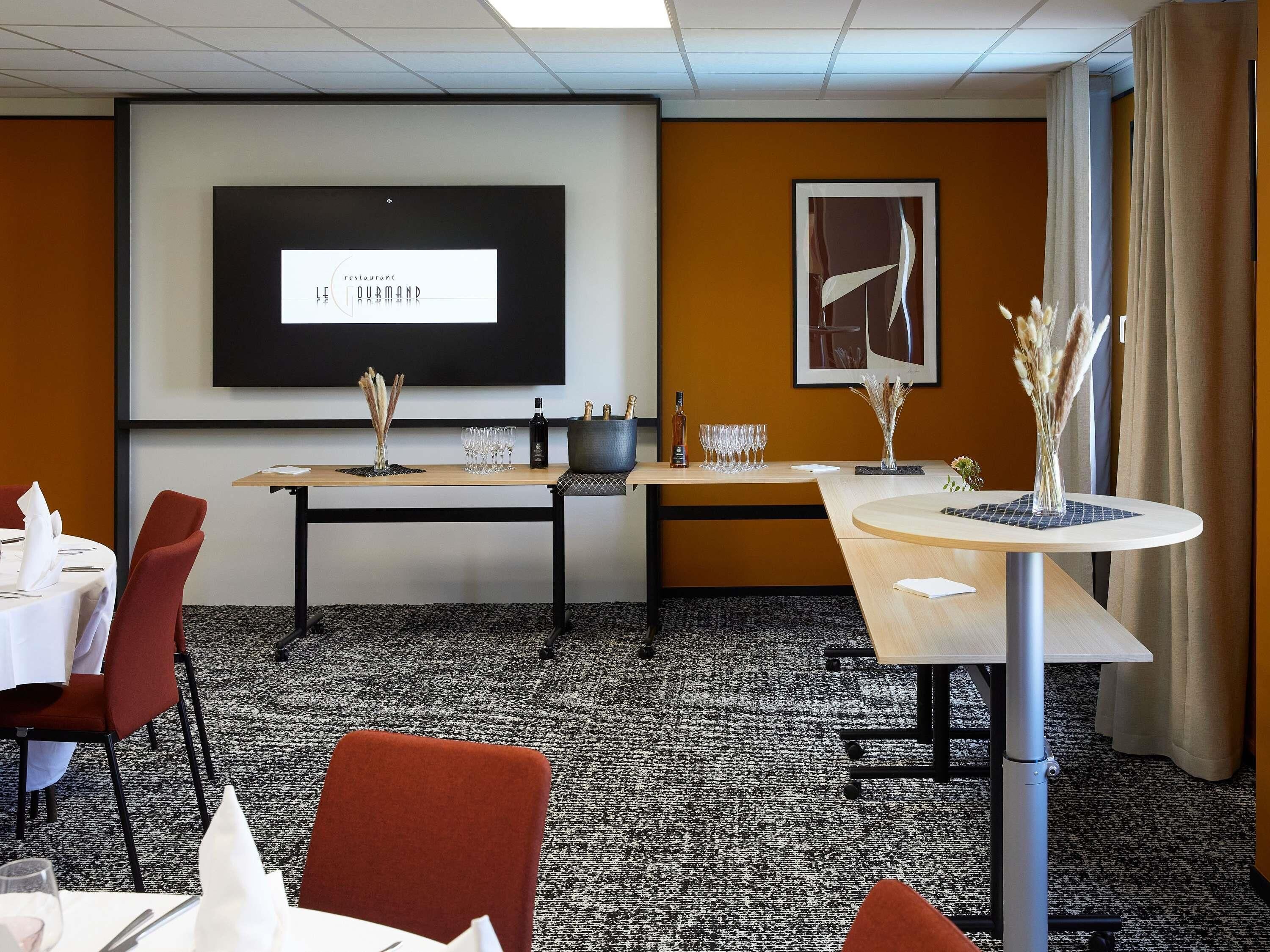 Une salle de conférence d’hôtel équipée d’un écran de projection, d’une table à manger et d’une petite table sur laquelle est posé un vase.