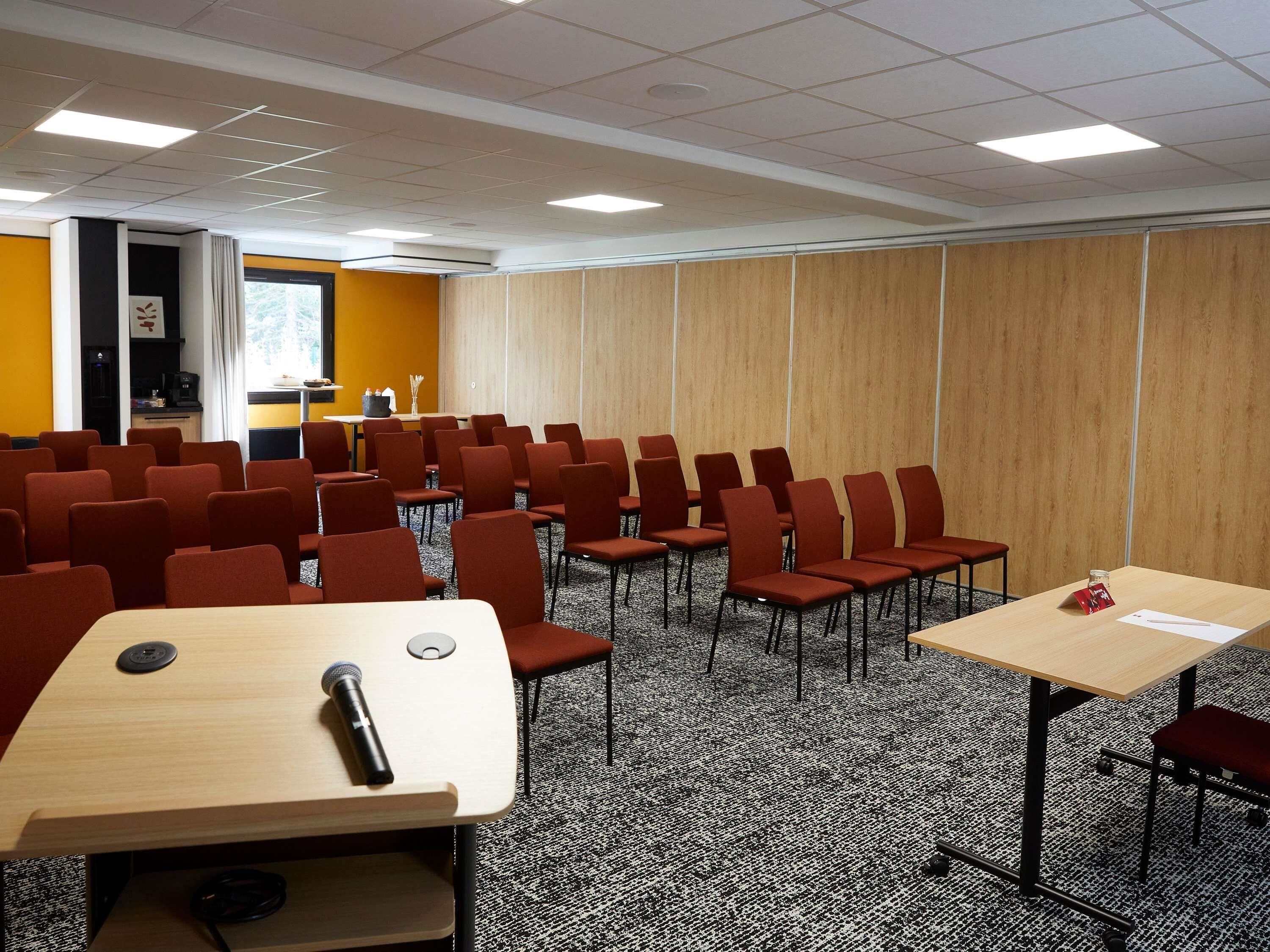 Una sala conferenze con file di sedie, un tavolo con un microfono e una piccola cucina sullo sfondo.