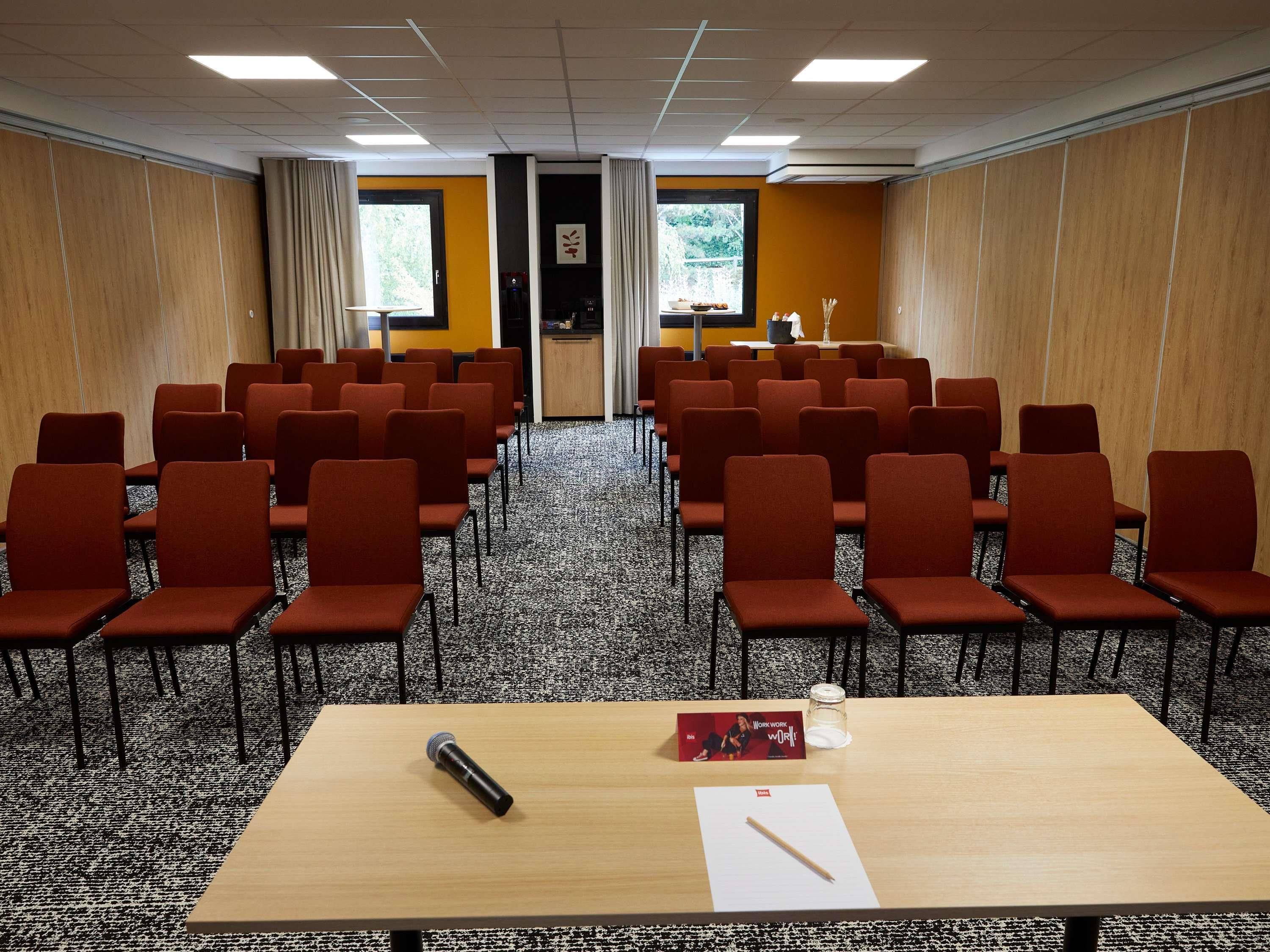 Un salón de conferencias con filas de sillas rojas, una mesa de madera, un micrófono, un libro y un vaso de agua.
