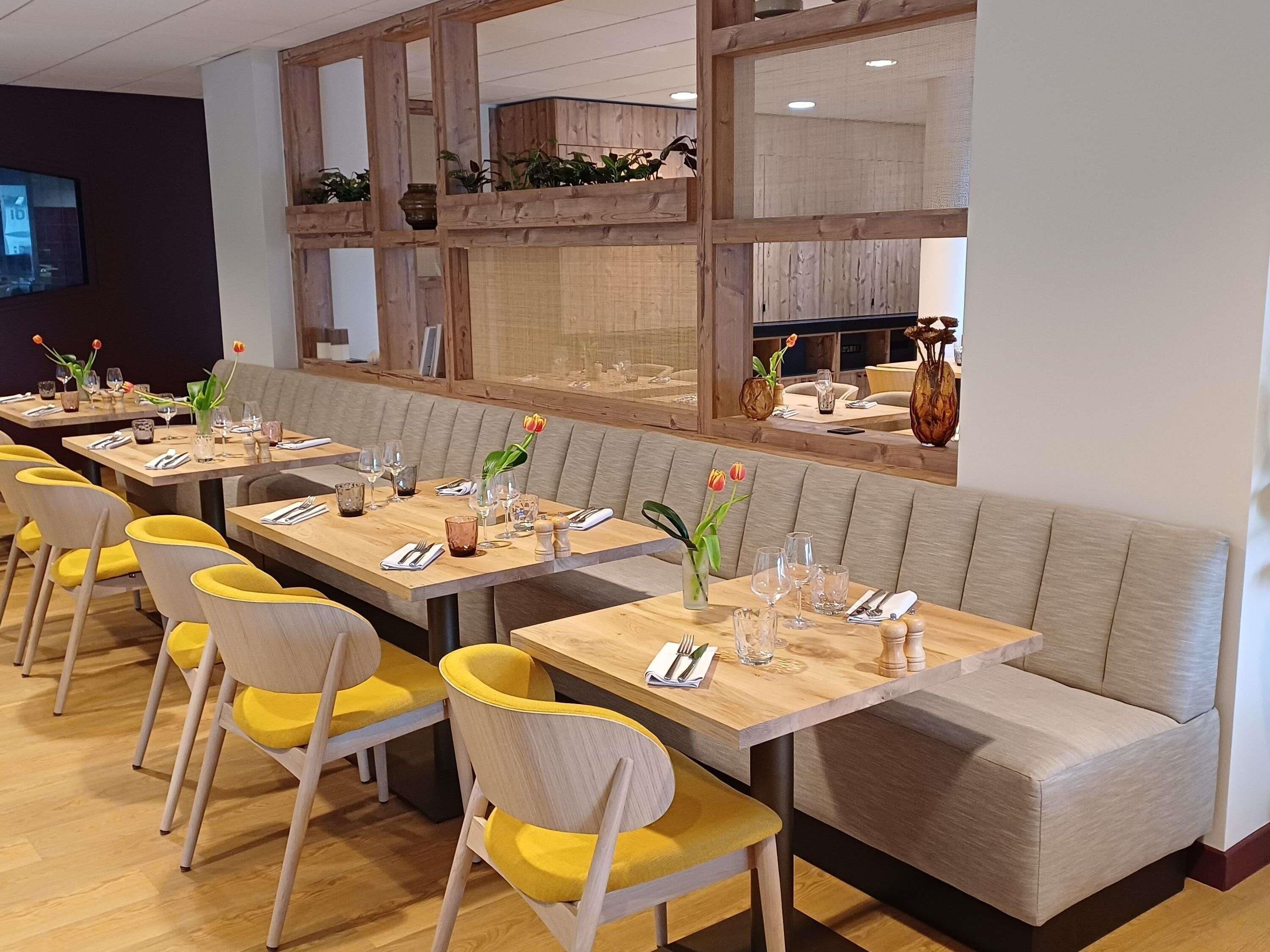 Interior de restaurante con varias mesas, sillas con cojines amarillos y un separador de madera con plantas.