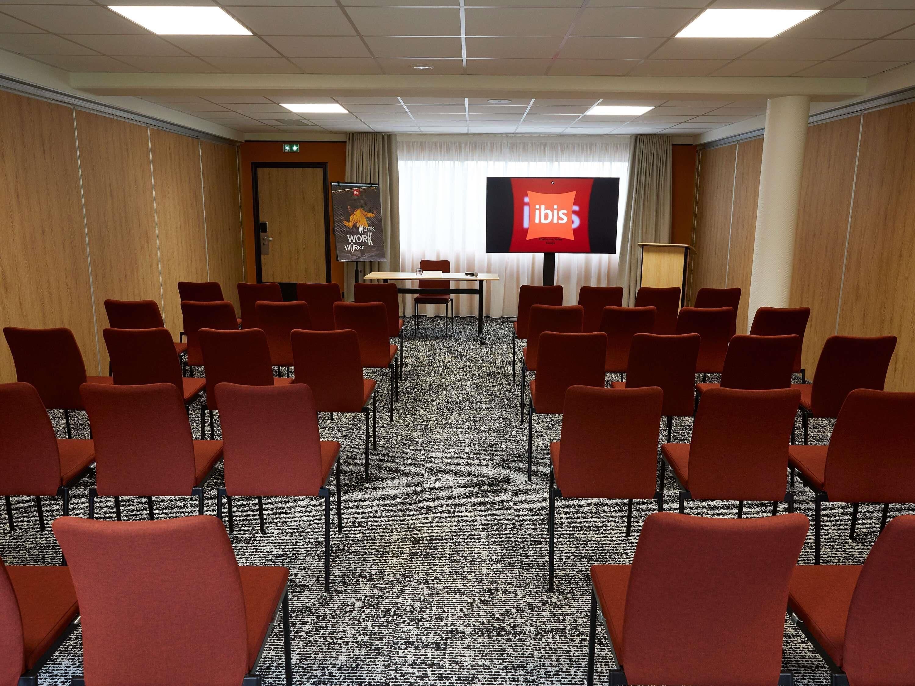 A conference room with rows of chairs, a table, and a large screen displaying the ibis logo.