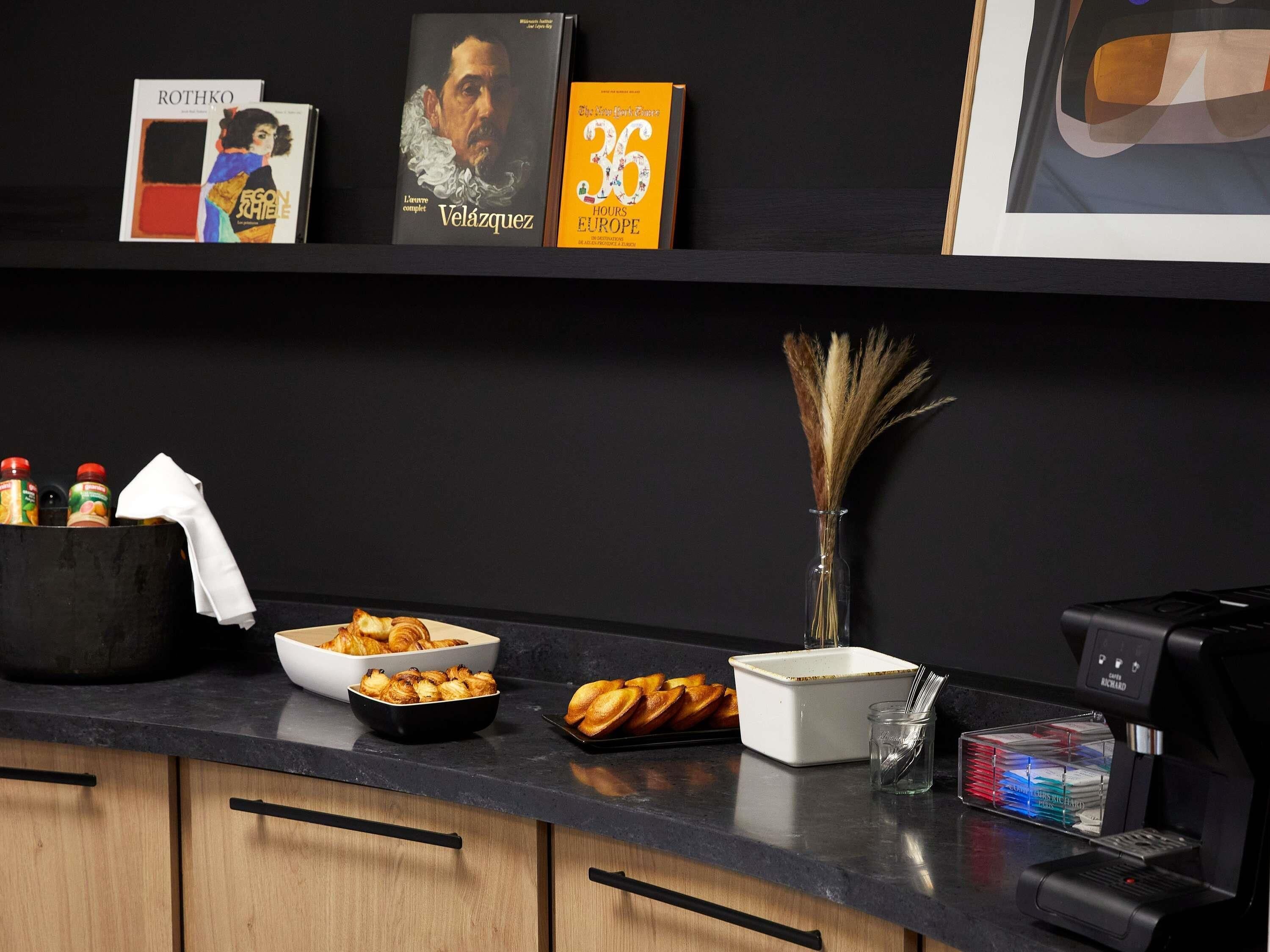 Een keukentafel met een koffieapparaat, een schaal croissants, een vaas met gedroogde planten en een boek getiteld 'Velázquez'.